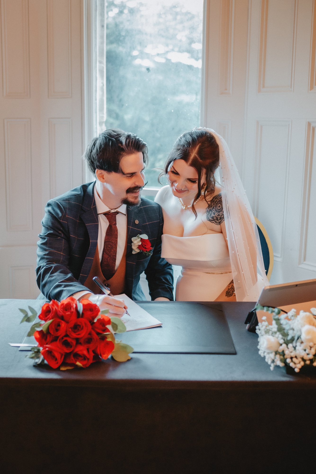 A couple signing a marriage certificate at a wedding ceremony, with a bouquet of red roses on the table.