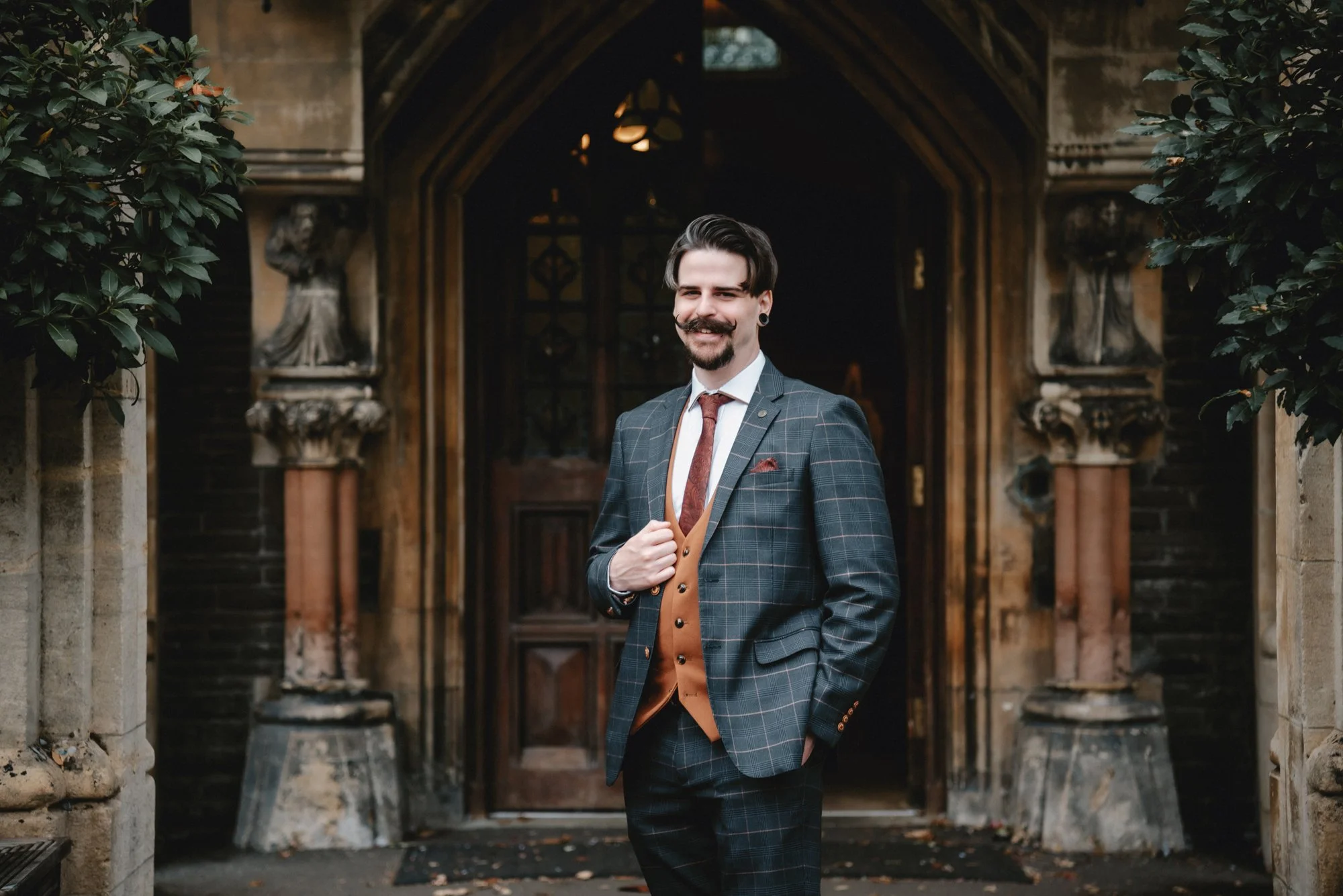A smiling man in a plaid suit standing outside a historic building with a wooden arched doorway and statues on the sides.
