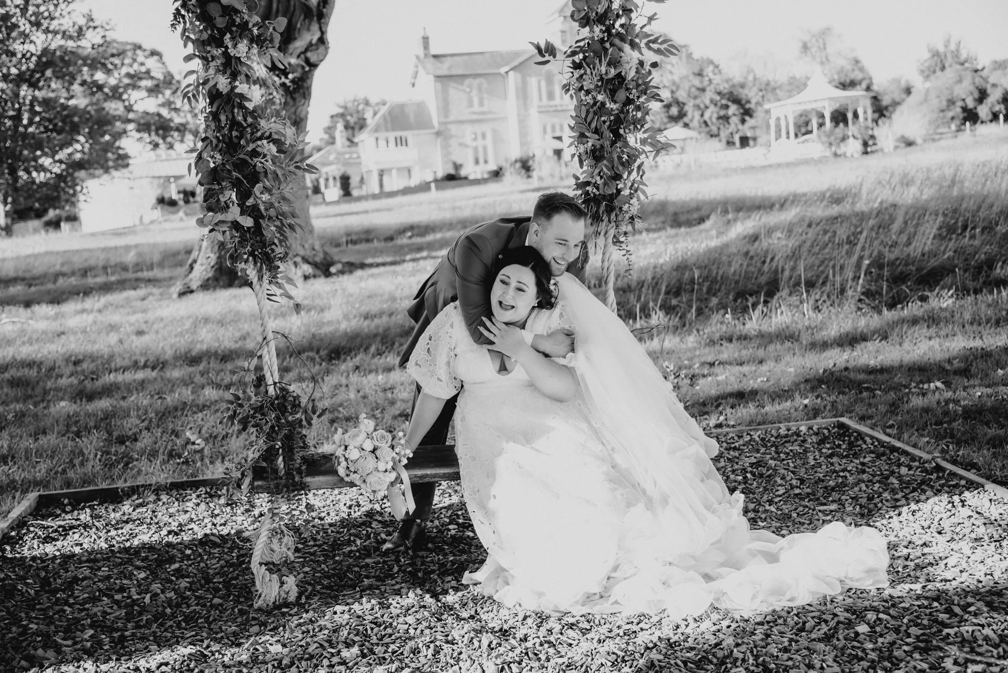A newlywed couple sitting on a bench under a decorated wooden archway outdoors, smiling and embracing each other on their wedding day.