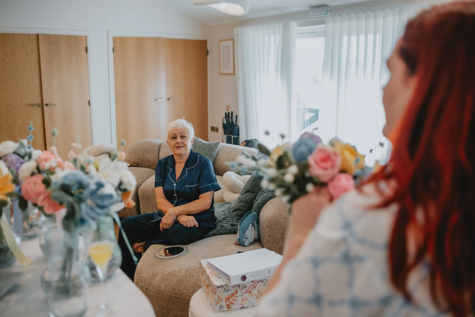 An elderly woman sitting on a beige couch, looking towards a young woman with red hair holding a bouquet of flowers, in a well-lit living room with white curtains and wooden cabinets.