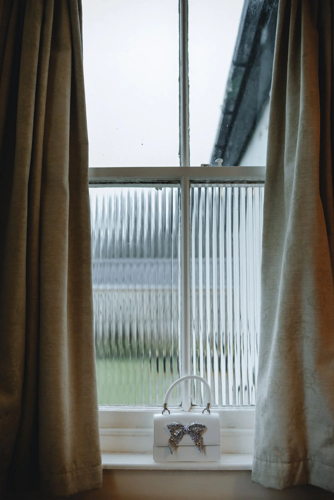 A window with beige curtains and frosted glass, with a white handbag featuring a silver bow on the windowsill.