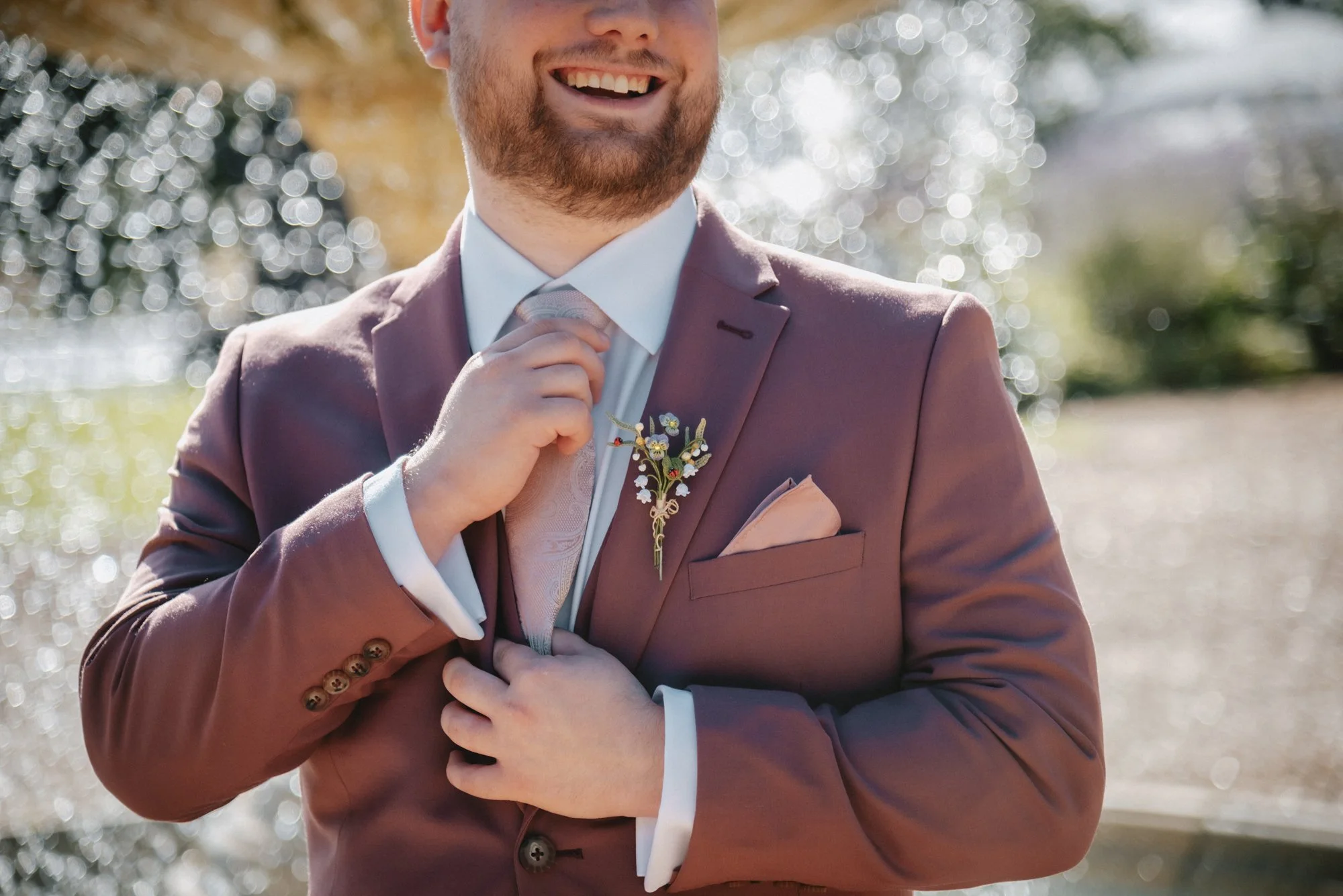 A man dressed in a mauve suit adjusting his tie outdoors with blurred trees and sunlight in the background.