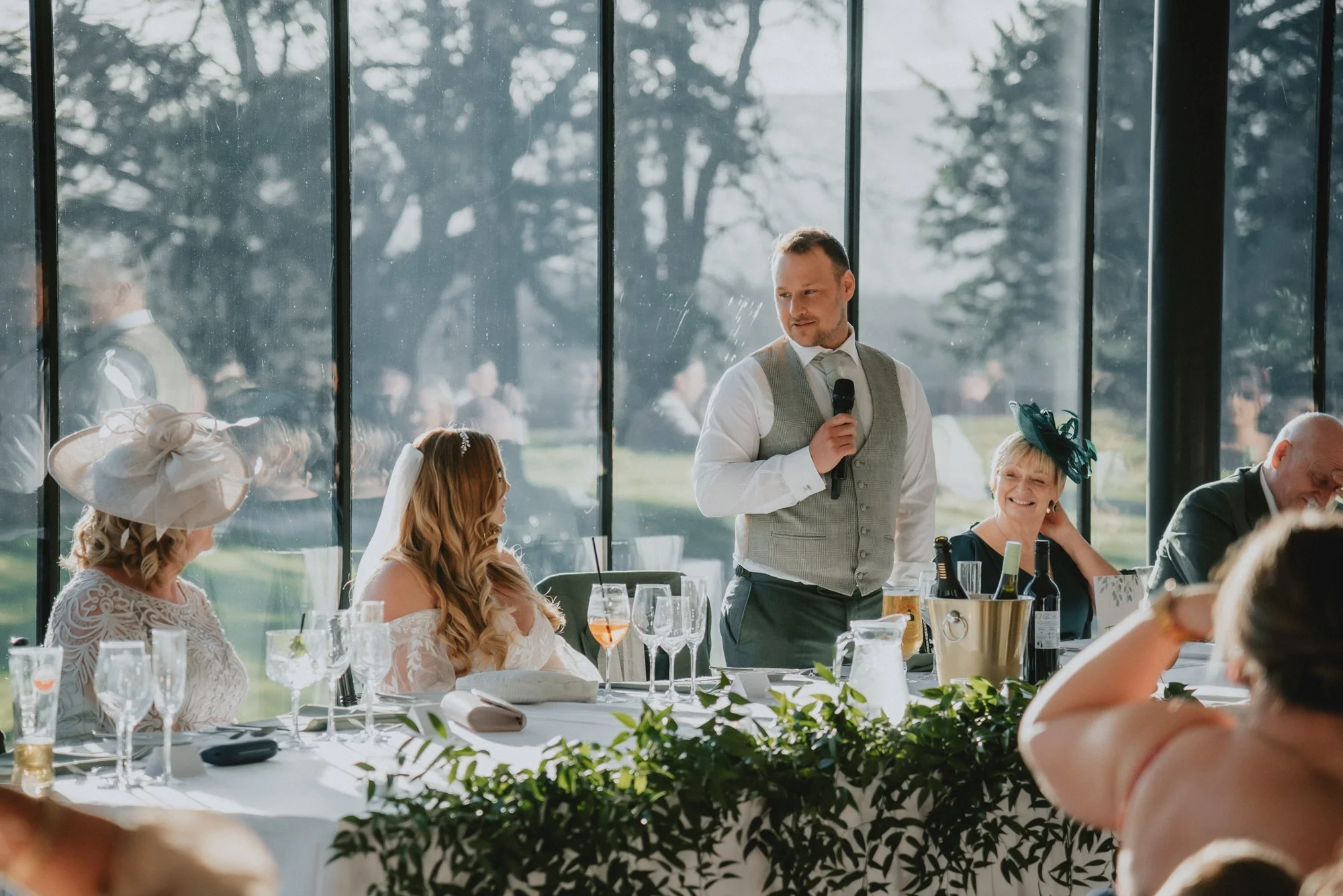 A man giving a speech with a microphone at a wedding reception, seated at a table with smiling guests, some wearing fancy hats and dresses, in a bright venue with large windows showing an outdoor view.