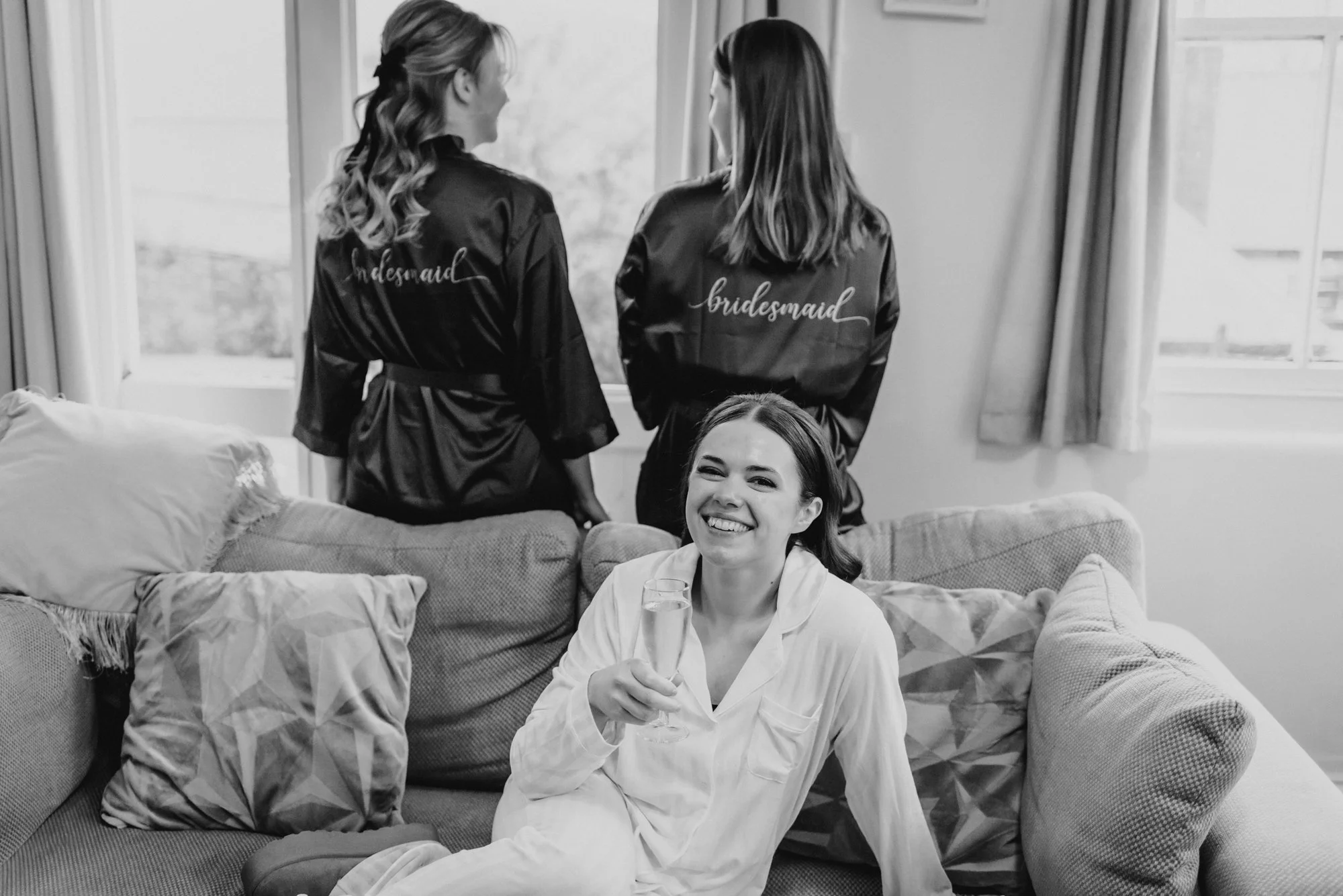 A woman in white pajamas sitting on a couch, smiling and holding a glass of champagne, with two women in robes with 'bridesmaid' and 'bride' embroidered on the back, standing by a window in a living room.