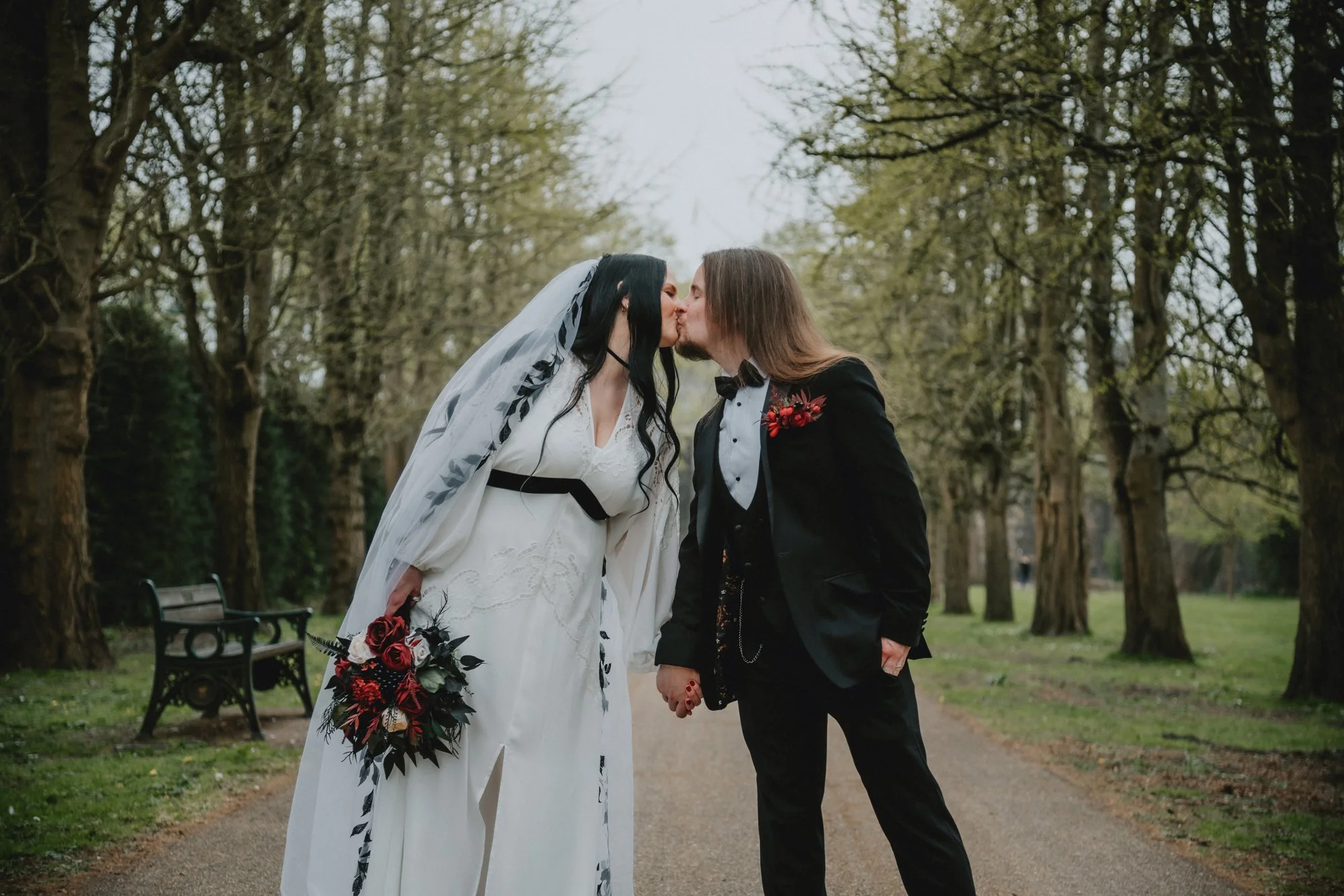 Two brides kissing in a park, with trees and benches in the background.