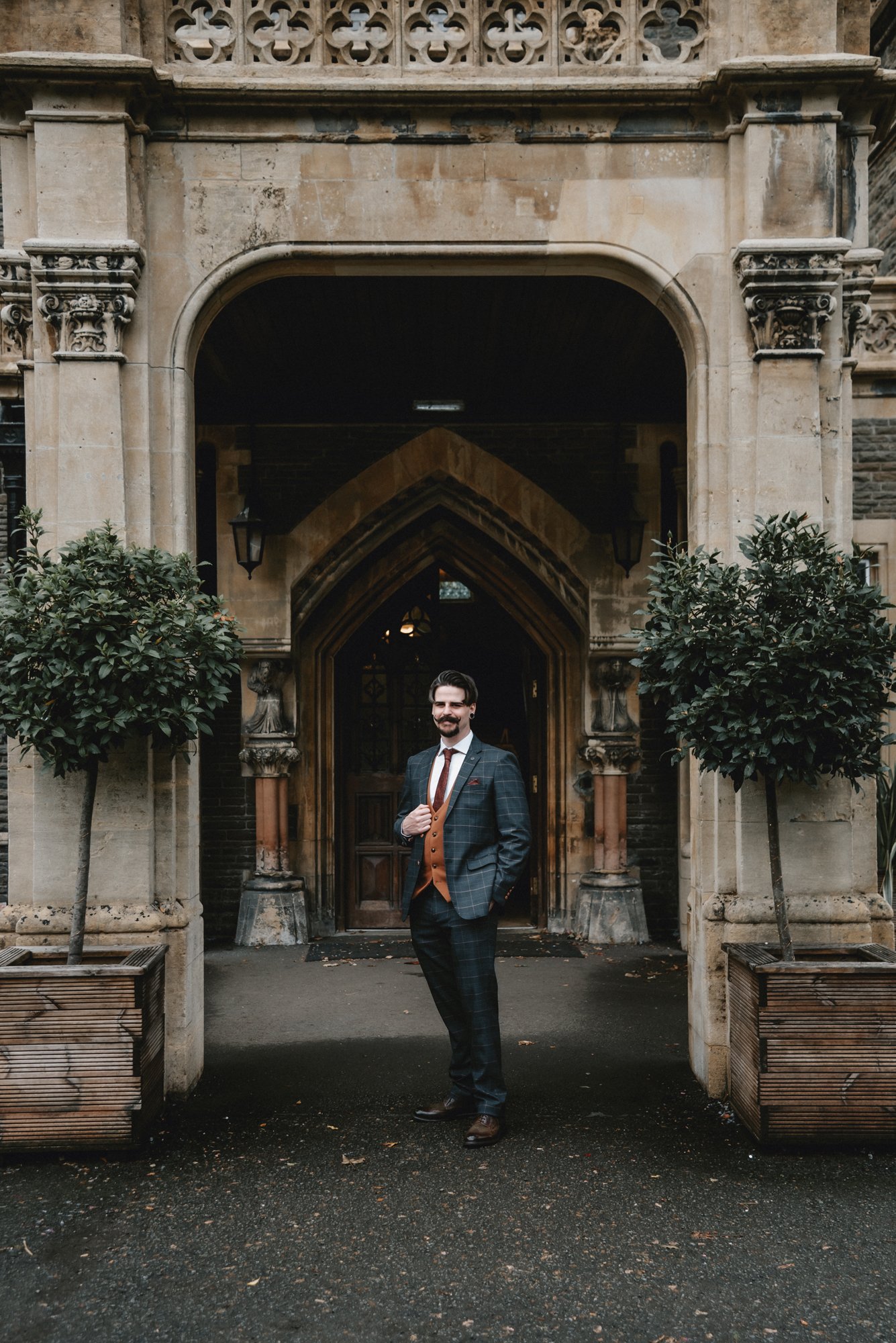A man in a plaid suit with a brown vest and tie stands outside an ornate stone building, smiling and adjusting his lapel.