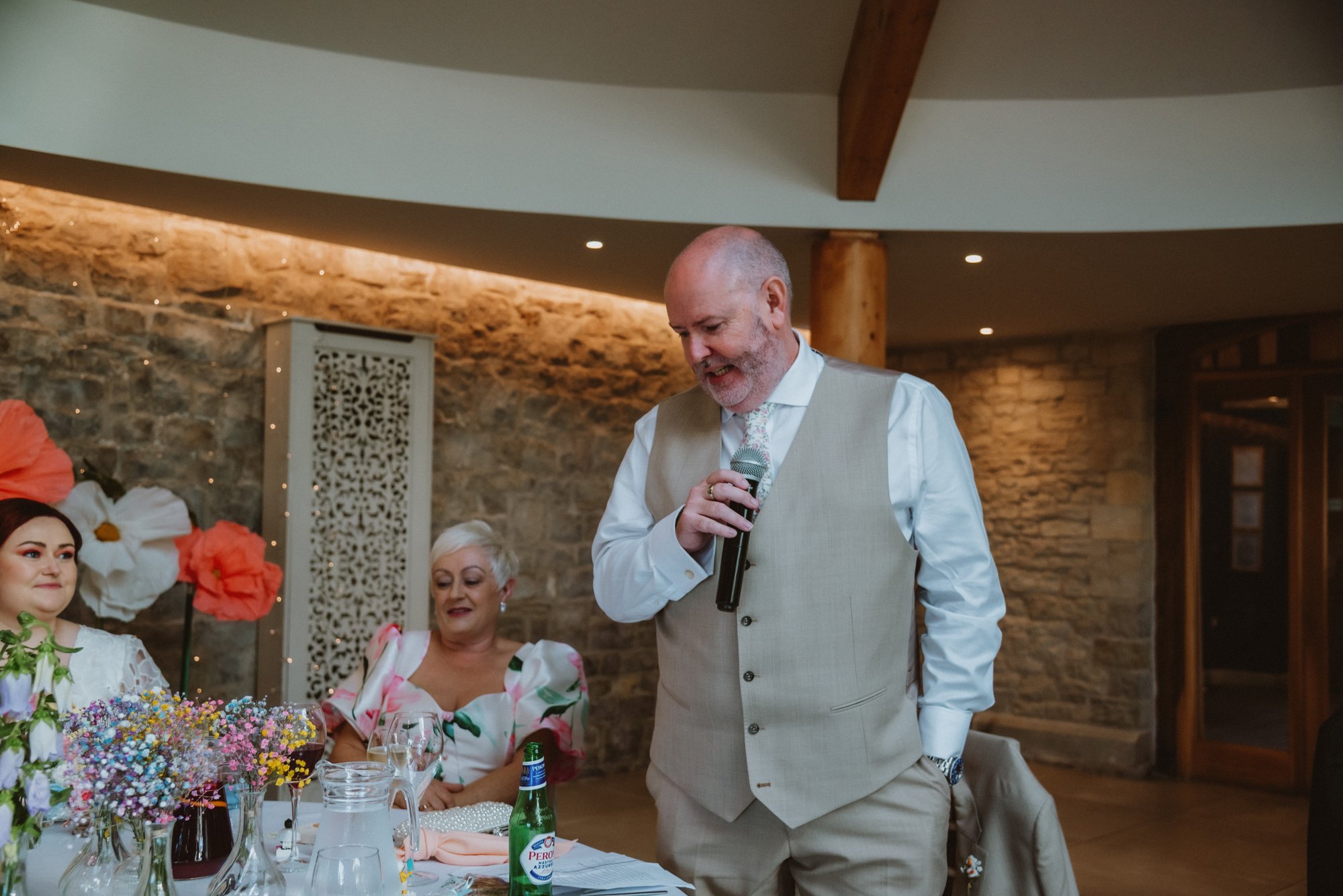 A man in a beige vest and white shirt giving a speech at a wedding reception, holding a microphone, with a woman in a floral dress and two women sitting at a table decorated with flowers in the background.