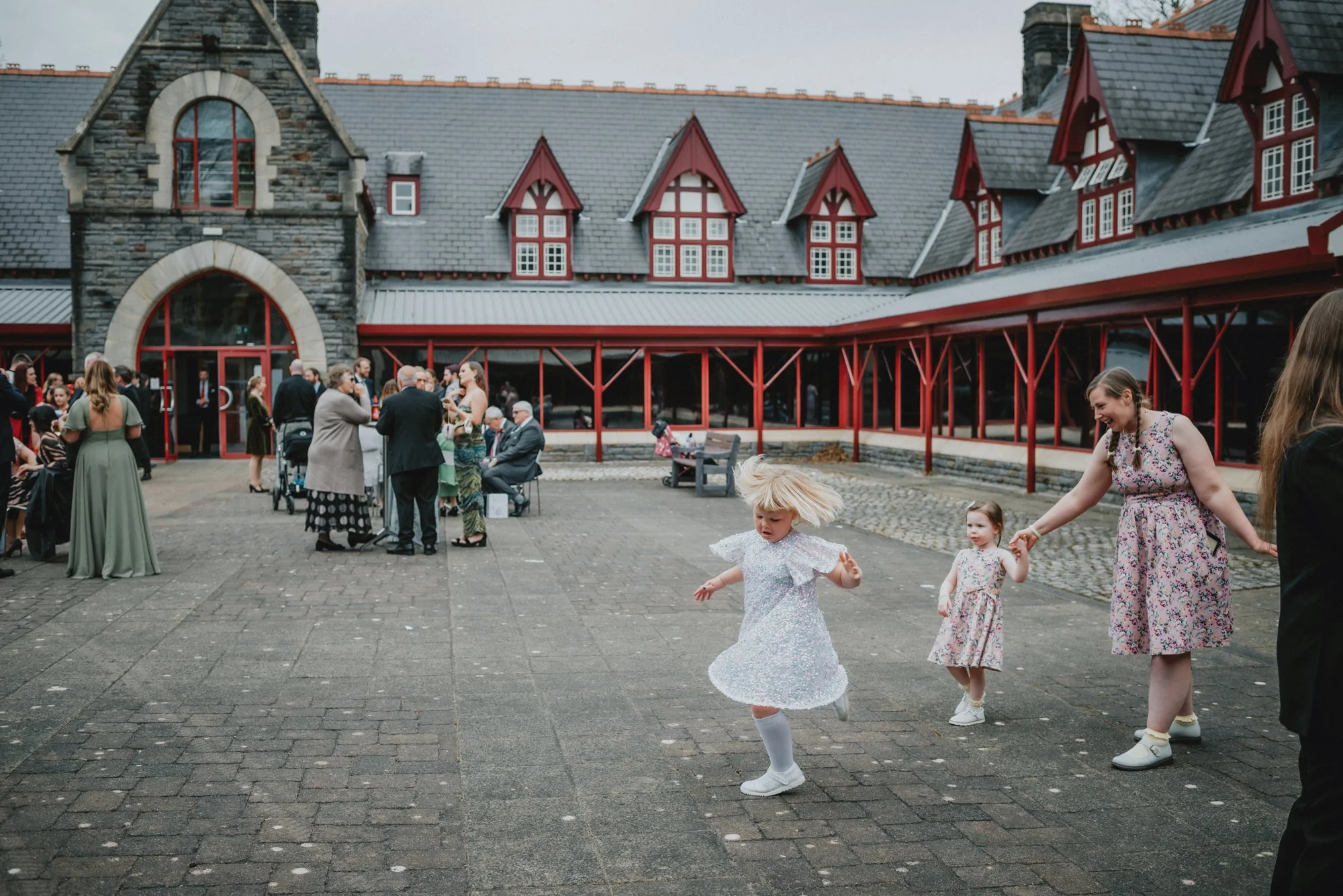 Children and adults gathered outside a stone and red brick building, with some of the children playing and a woman holding one child's hand.