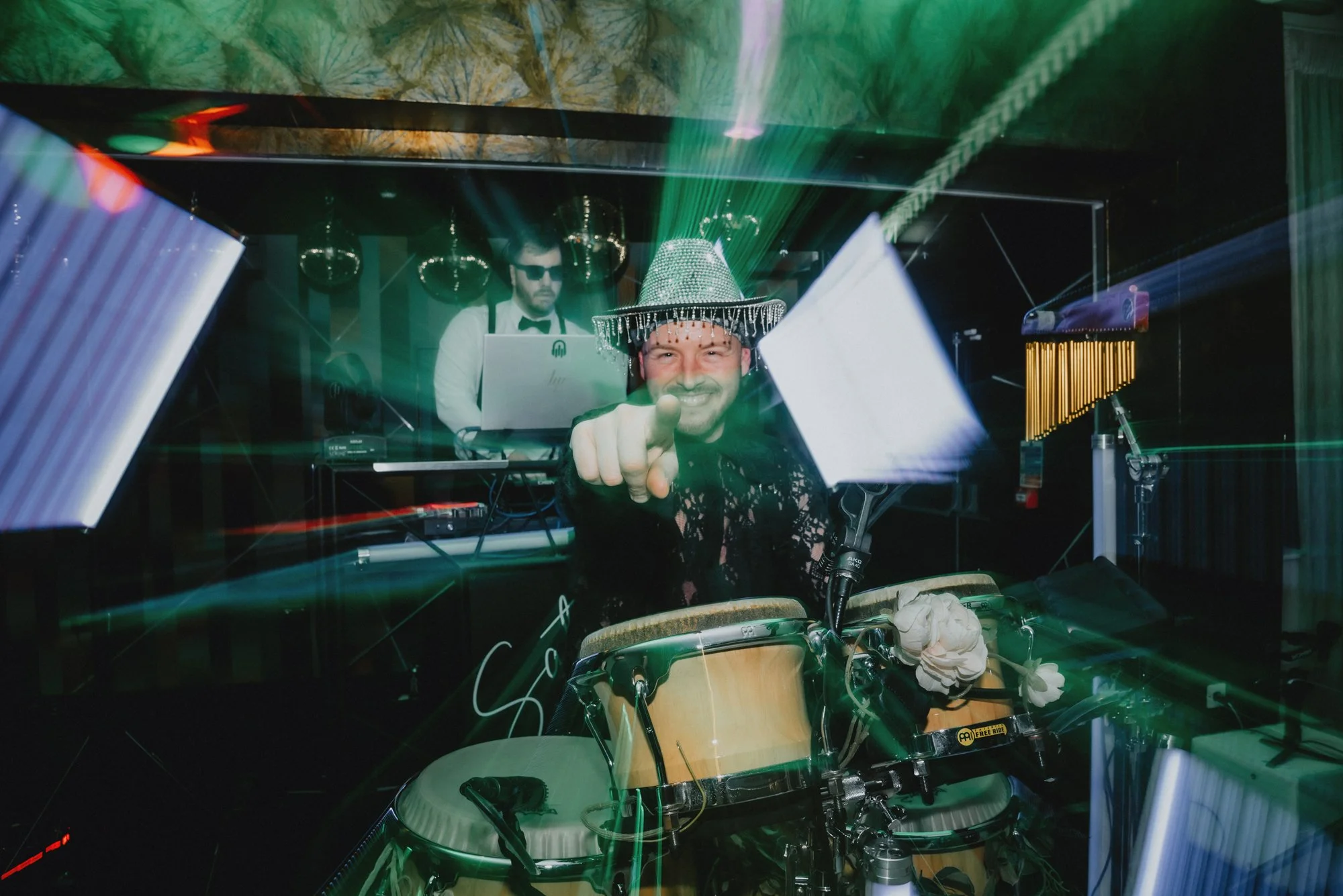 DJ wearing a shiny silver hat and black lace outfit, smiling and pointing at the camera while playing conga drums. A DJ in sunglasses and a white shirt with a black bow tie is working on a laptop in the background, with colorful lights and disco ball