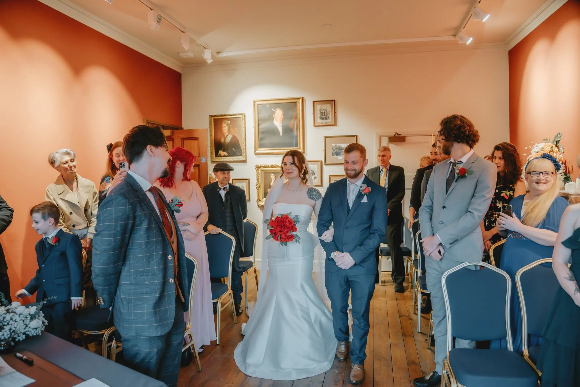 A wedding ceremony with bride and groom walking down the aisle surrounded by friends and family in a decorated room.