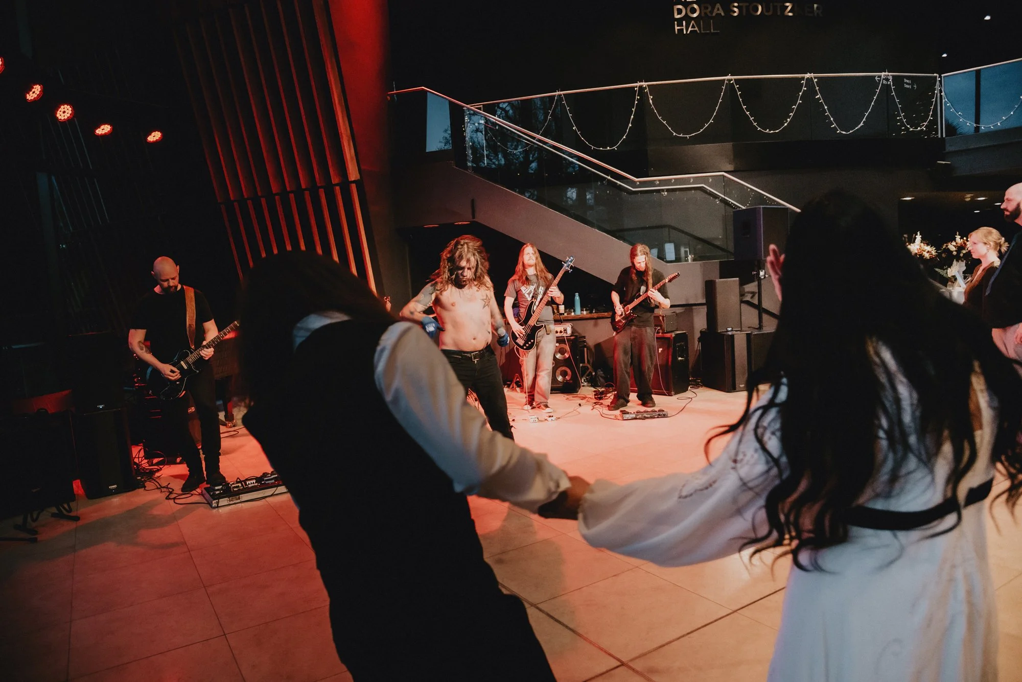 People dancing in front of a band performing on stage in a dimly lit indoor venue with string lights hanging overhead.