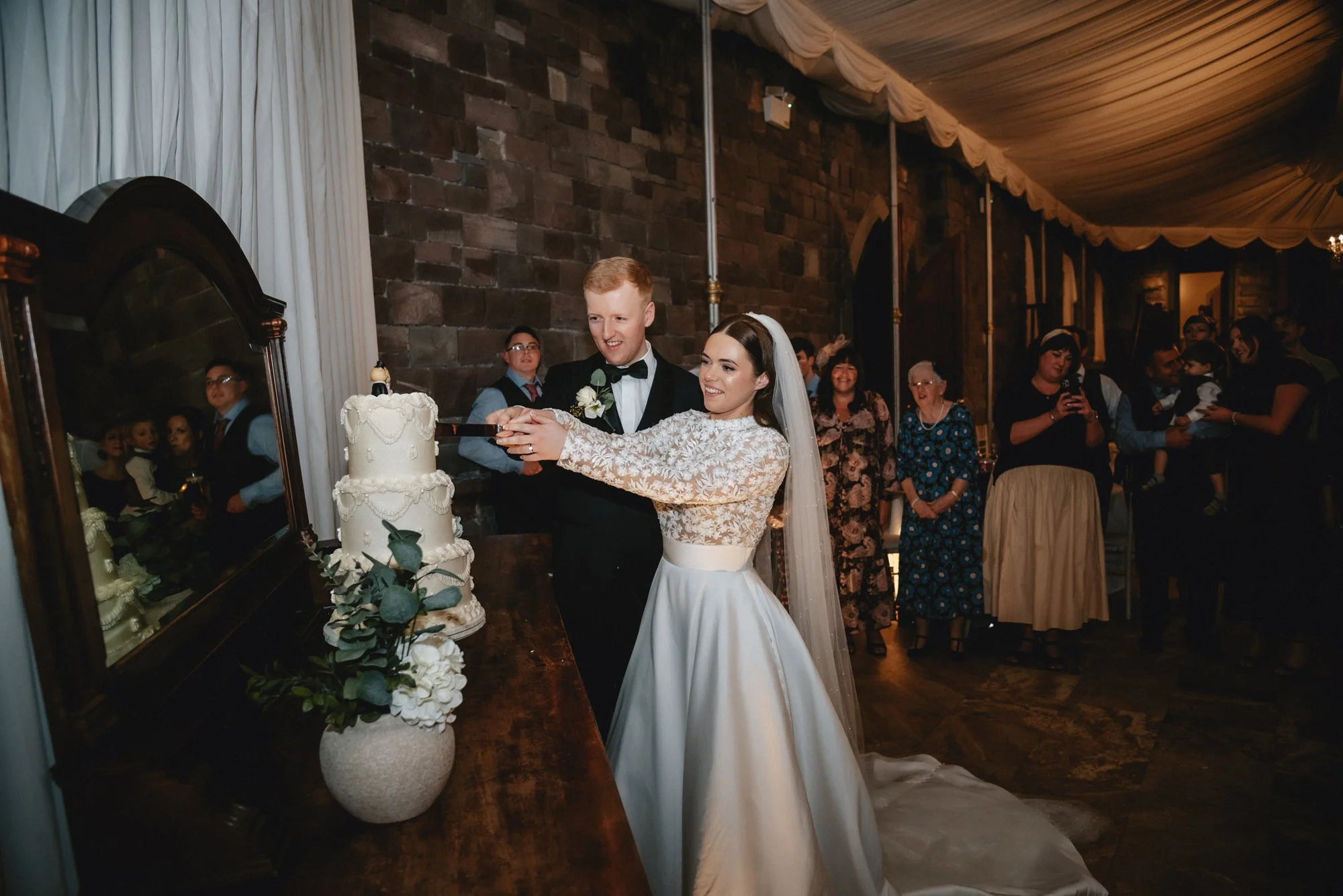 Bride and groom cutting a wedding cake together, standing near a mirror, with guests watching in the background.