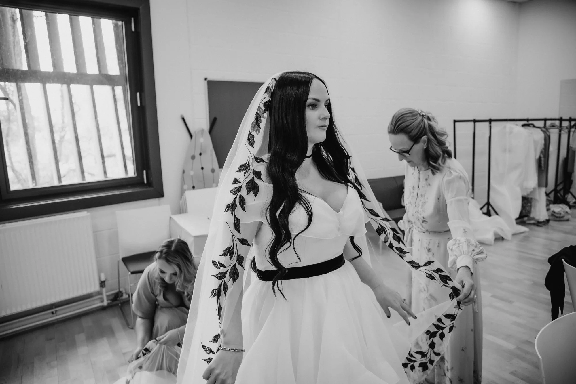 A bride with long dark hair in a wedding dress with floral embroidery on the sleeves is getting ready in a dressing room, with two women assisting her, one adjusting her dress and another kneeling on the floor.