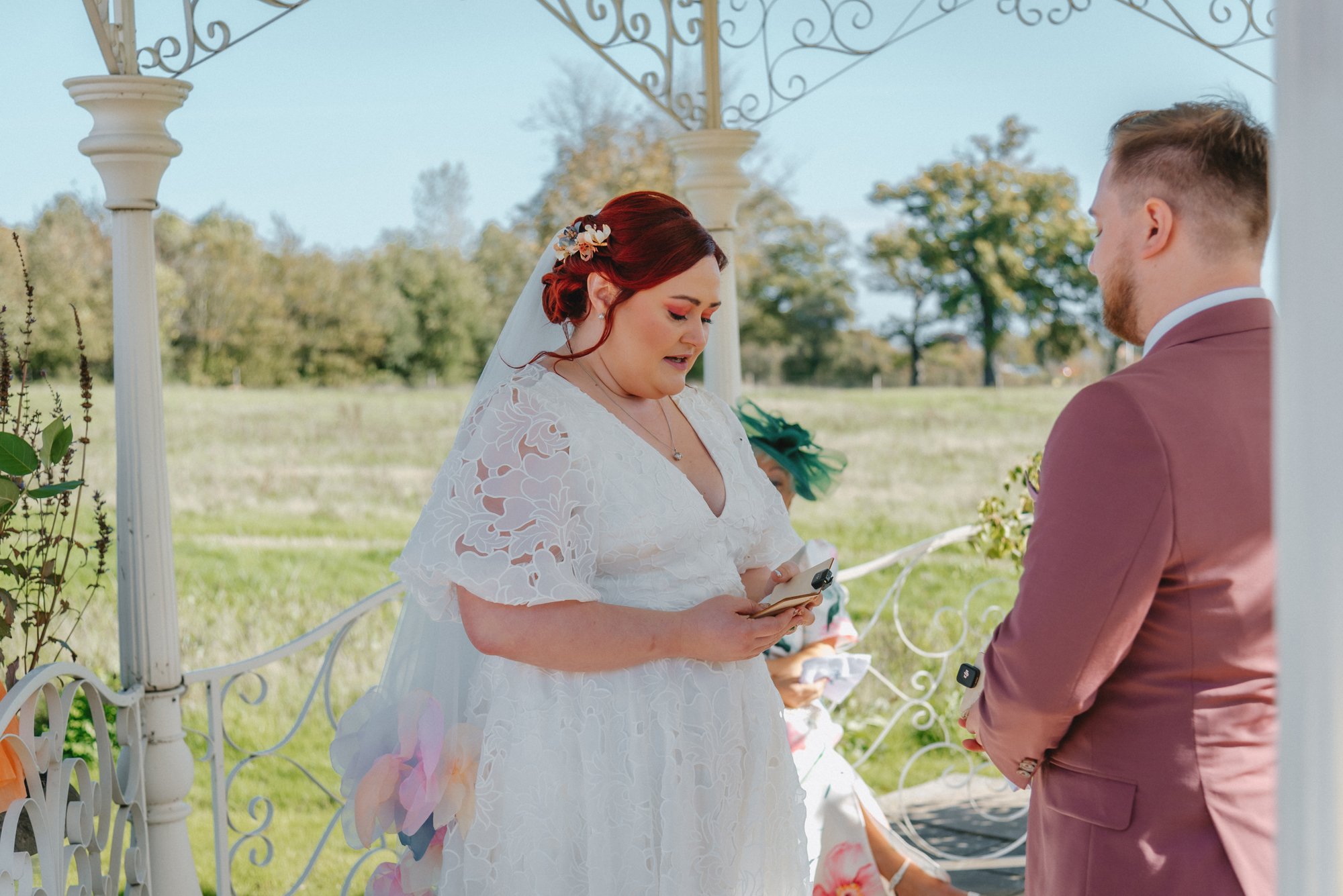 A bride and groom exchanging vows outdoors under a white decorative arch on a sunny day with trees in the background. The bride has red hair and is wearing a white lace dress with floral accessories in her hair. The groom is dressed in a pinkish-brow