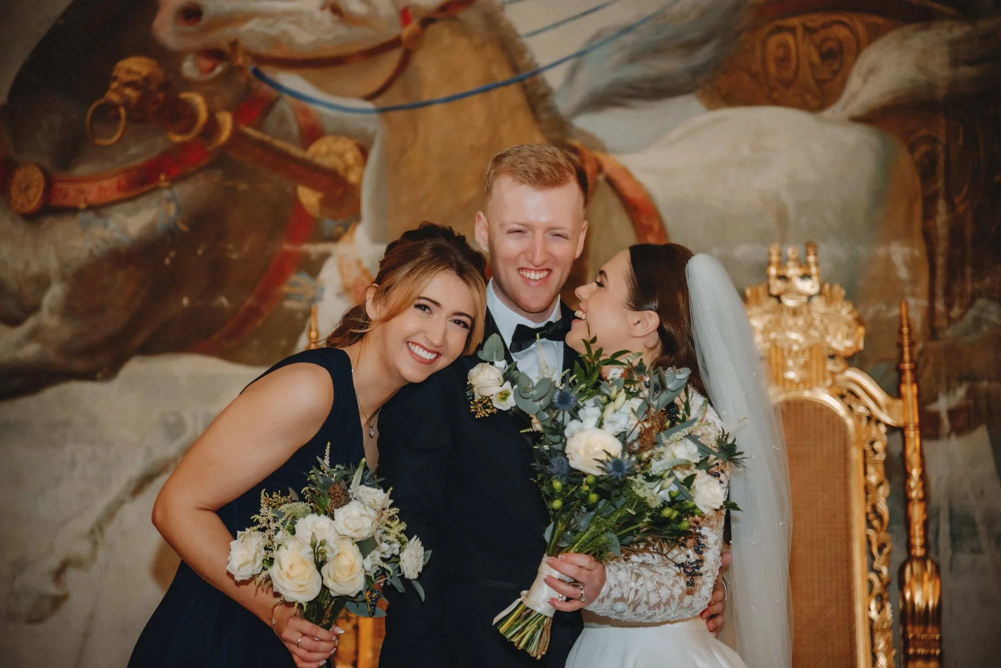 A group of four wedding guests, including a bride and groom, smiling and posing for a photo at a wedding reception, with a painted mural in the background.