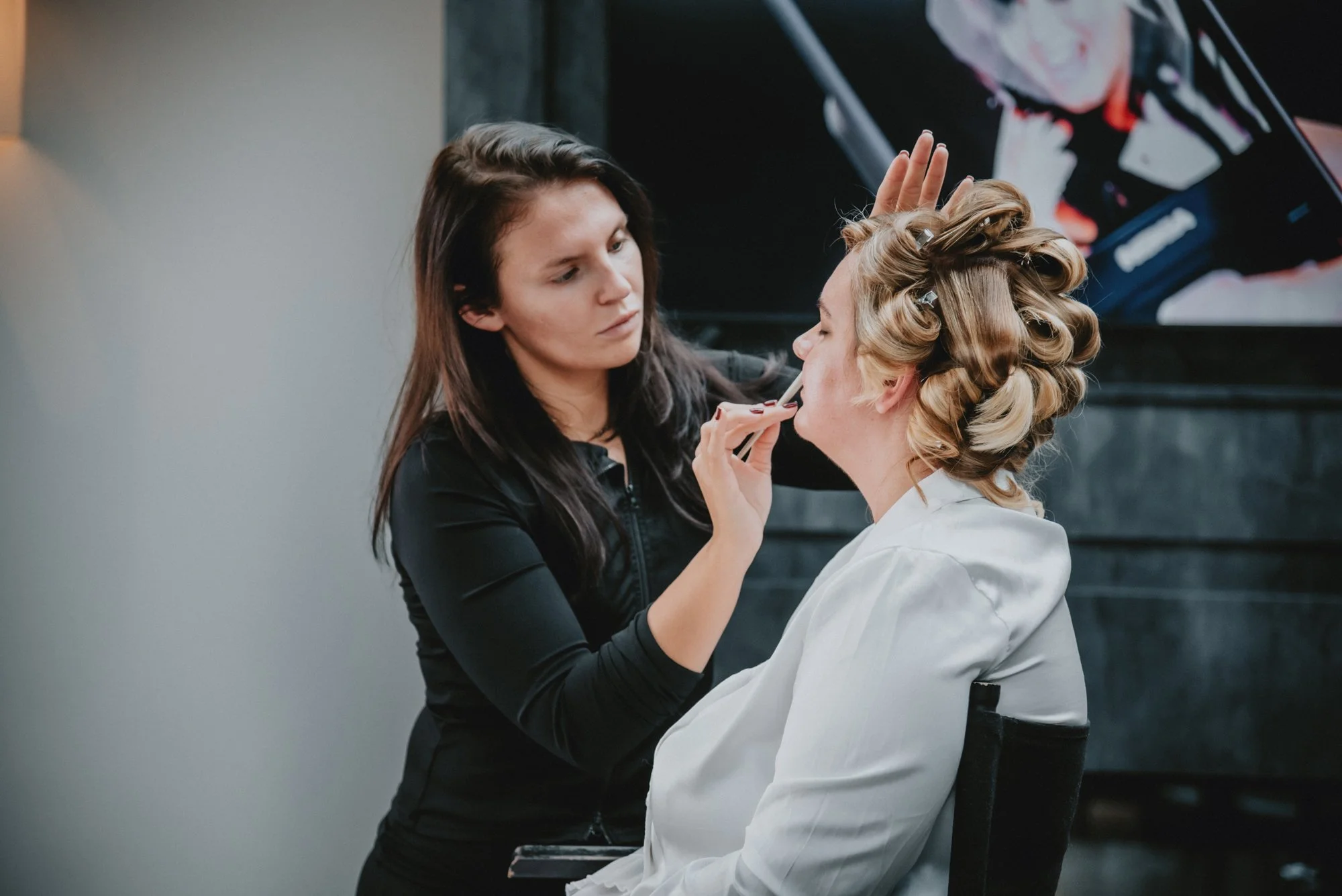 Makeup artist applying lipstick to a woman with curled hair sitting in a chair.