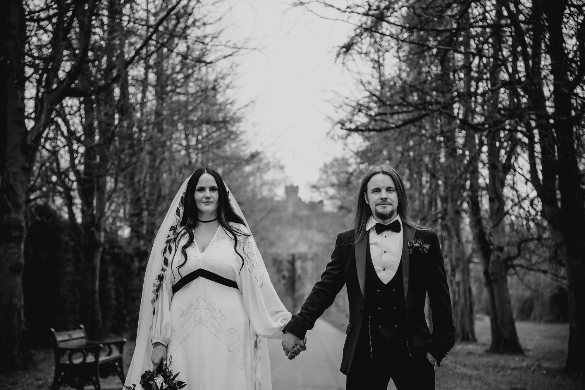 Black and white photo of a bride and groom holding hands while walking outdoors on a tree-lined path, dressed in wedding attire.