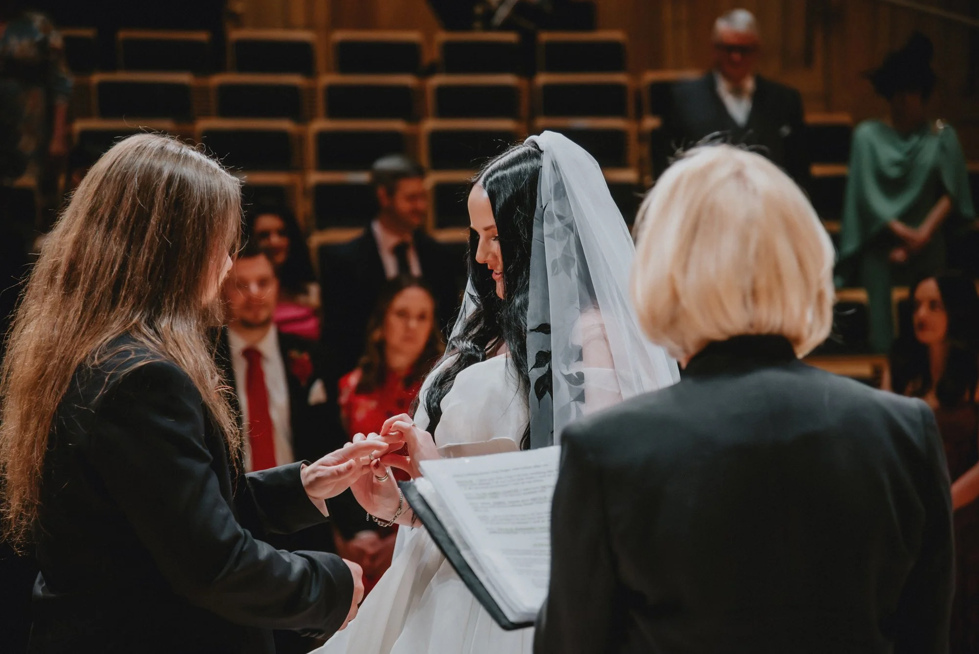 A wedding ceremony with a bride exchanging rings with her partner, officiated by a woman holding a book, with guests watching in the background.