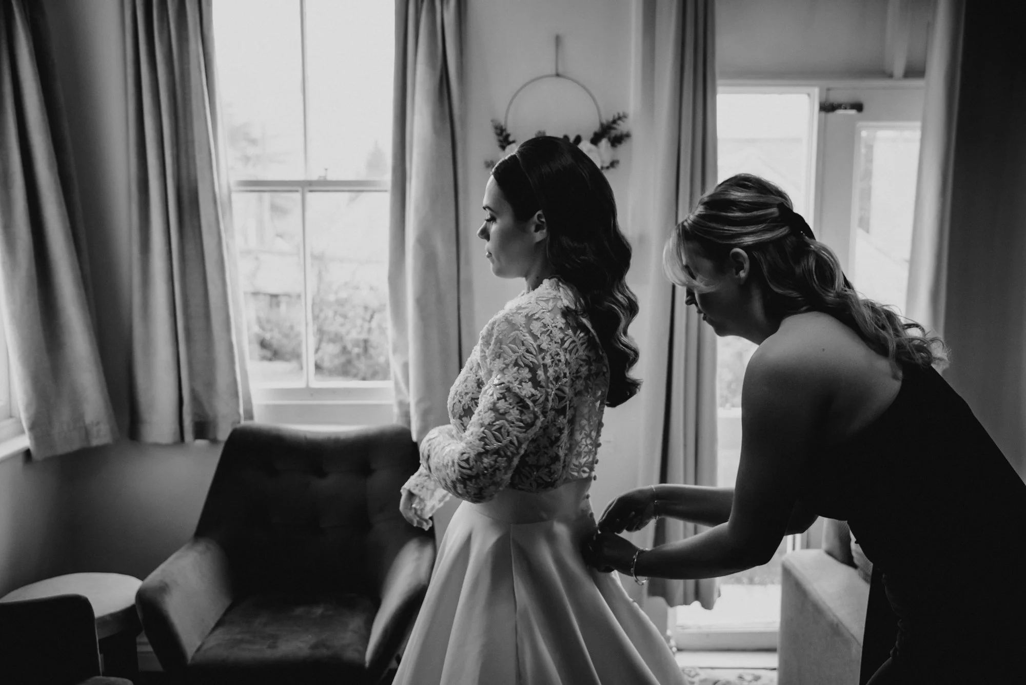 A bride in a lace gown standing in a room with her back to a woman, possibly helping her with her dress.