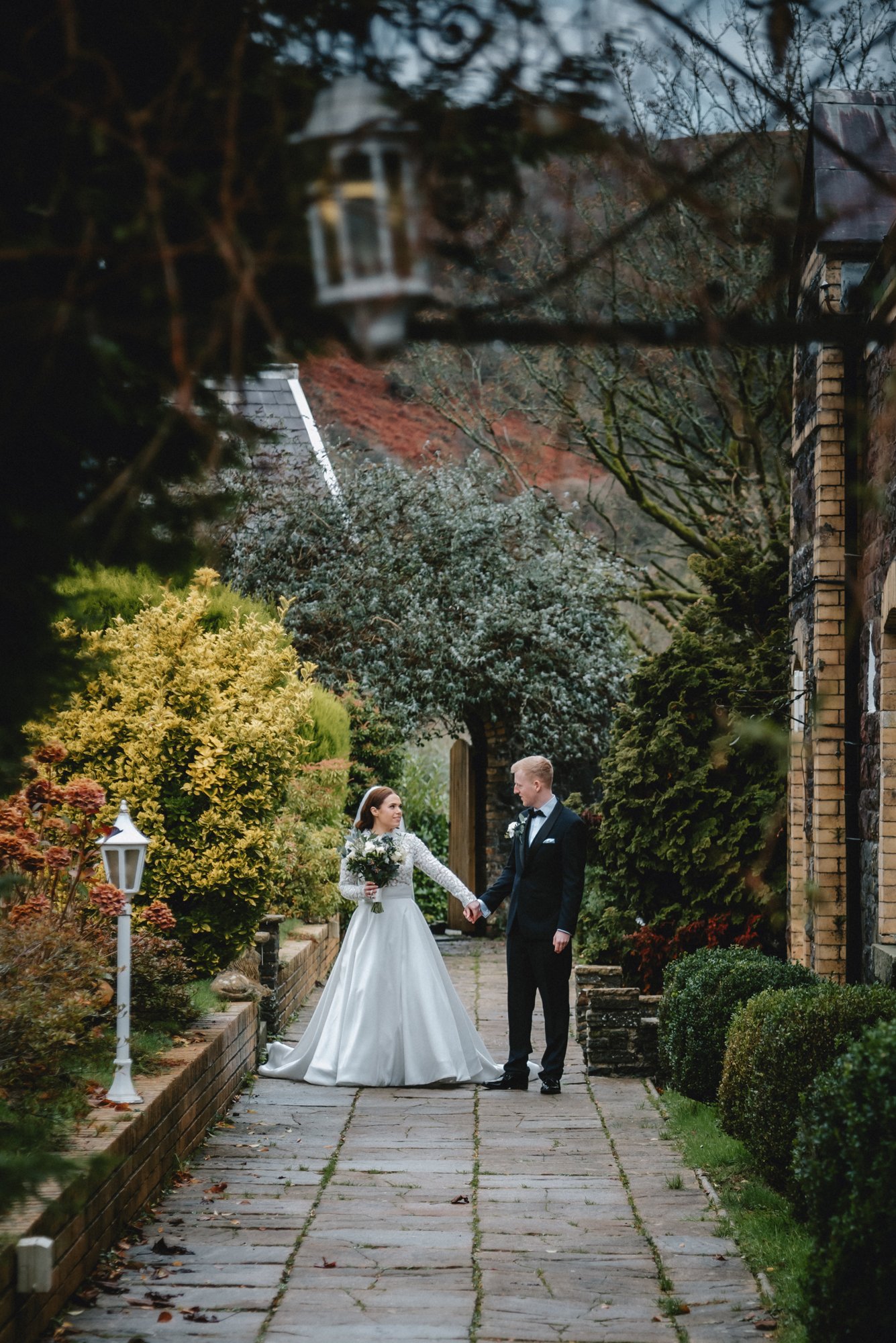 A bride and groom holding hands on a stone pathway surrounded by lush greenery and trees, during their wedding day.