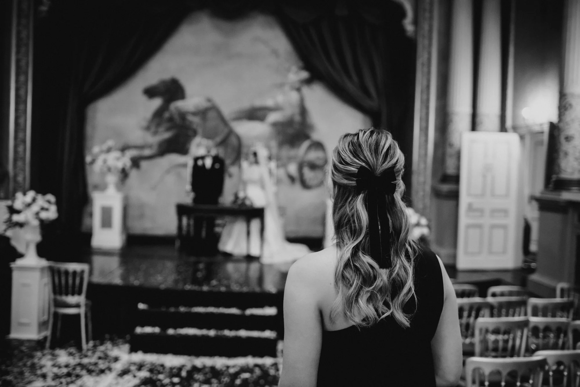 A woman with styled wavy hair tied with a black ribbon stands facing a stage, where a wedding ceremony is occurring in an ornate, classical room with large curtains and floral arrangements.