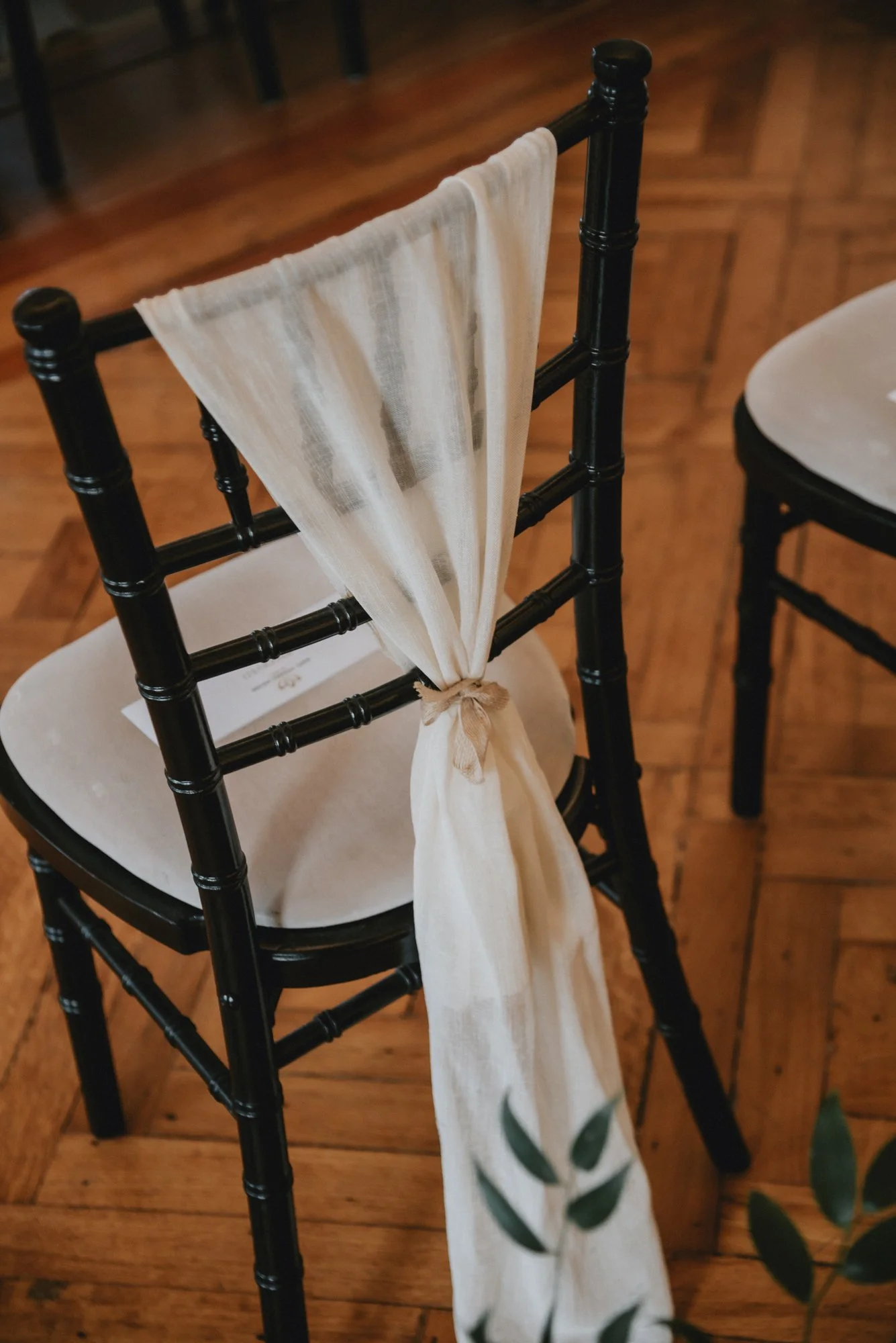 A black wooden chair with a beige cloth tied to the backrest as decoration, placed on a wooden floor.