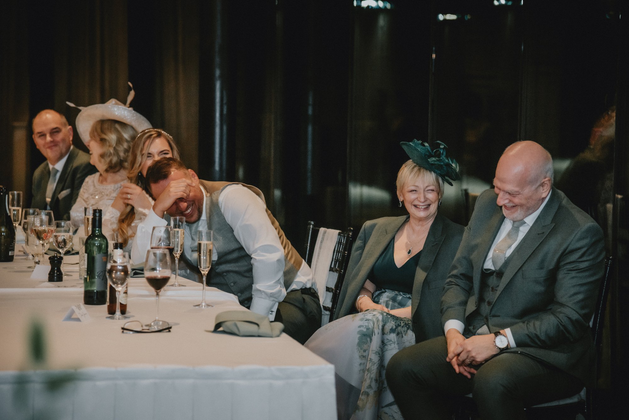 People sitting at a formal table, laughing and smiling during a celebration or event, with glasses and bottles on the table.