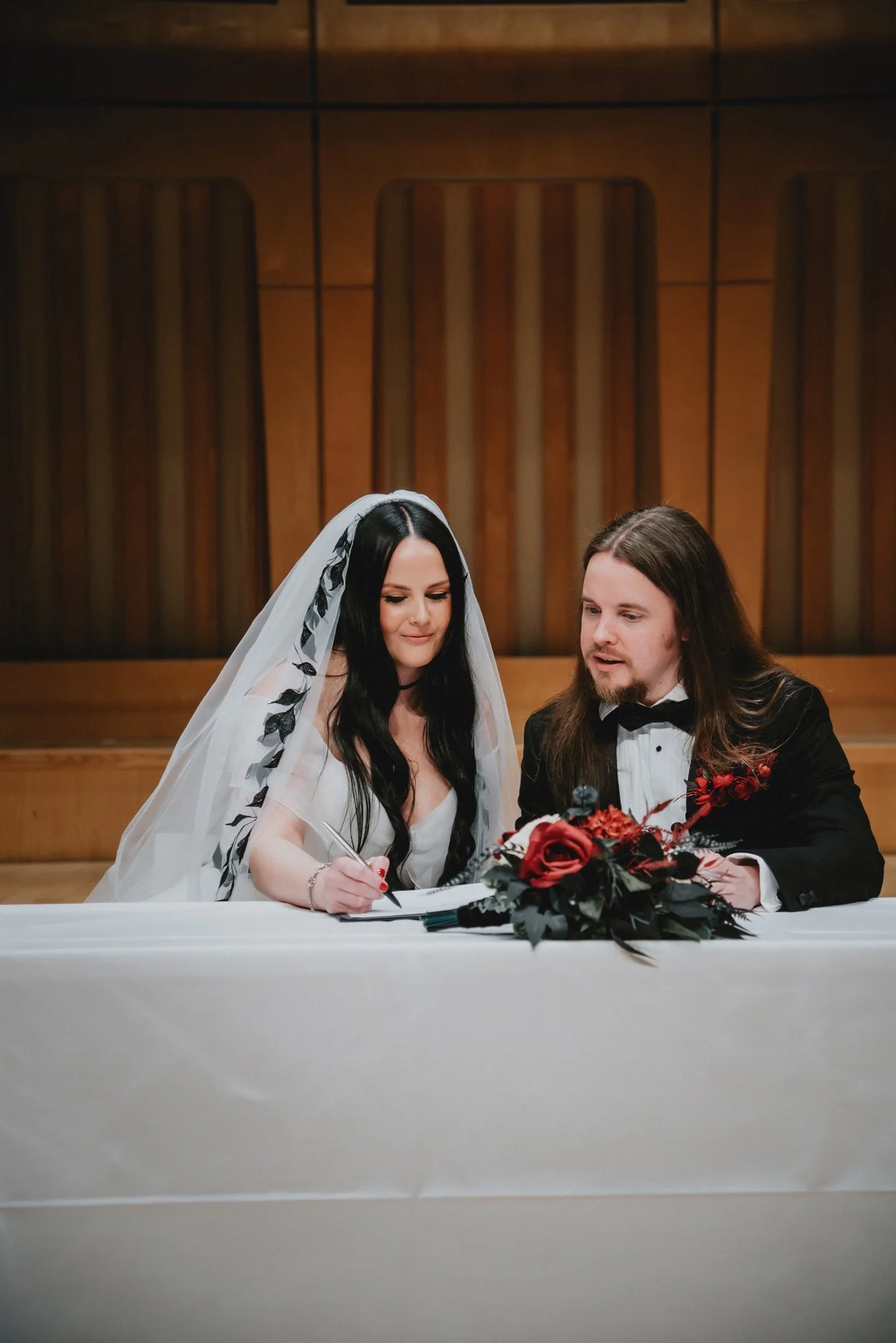 A bride and groom sitting at a table during their wedding ceremony, with the bride signing a document and the groom looking on. The bride has long black hair and is wearing a white gown with a veil, and the groom has long brown hair and is dressed in