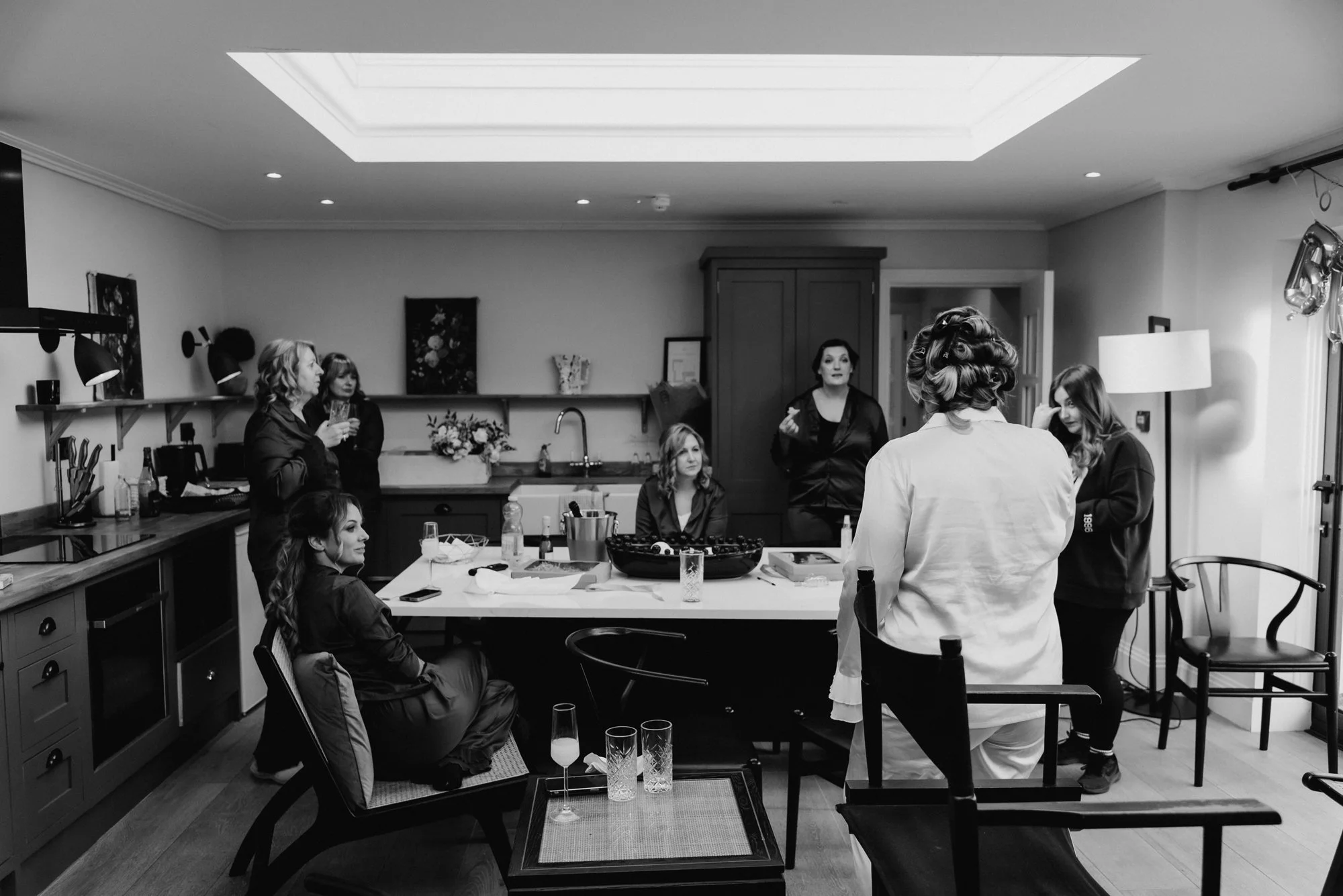 Women gathered in a kitchen, some sitting and some standing, engaging in conversation, with a woman in a robe with styled hair at the center, possibly preparing for an event or photoshoot.