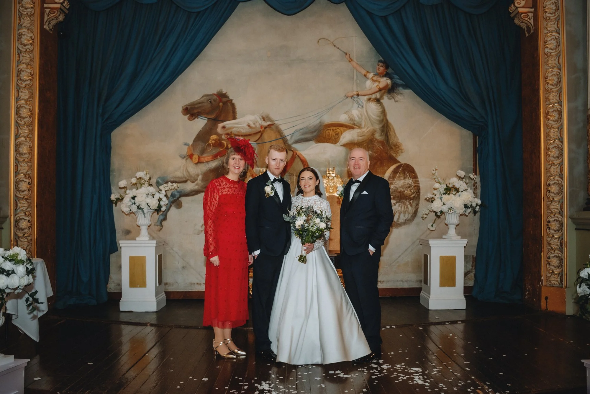 A wedding reception with four people standing in front of a large classical painting of a chariot scene. The bride is holding a bouquet of white flowers, flanked by three people in formal attire. The background features blue curtains, floral arrangem