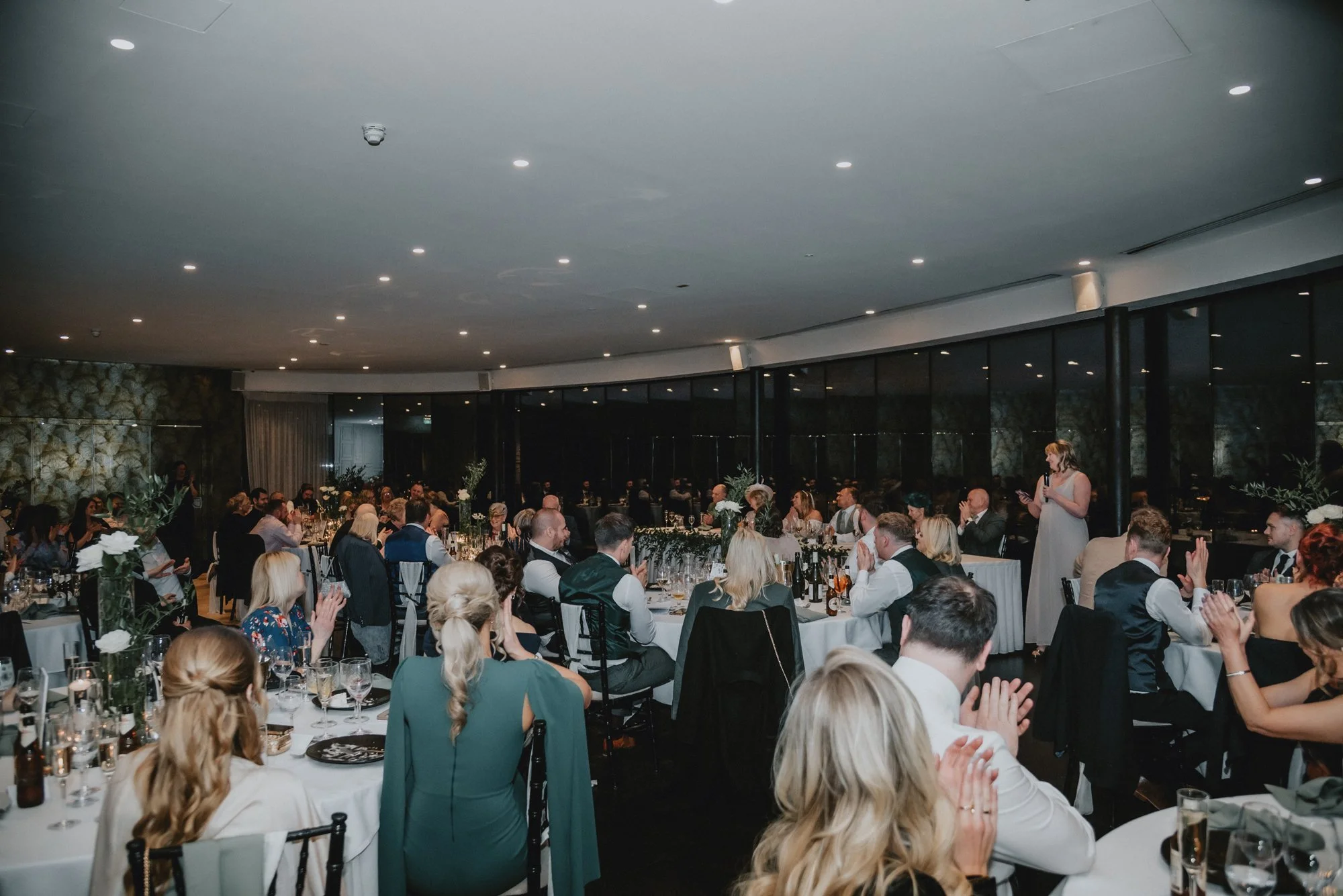 Guests seated at round tables in a banquet hall listening to a woman giving a speech