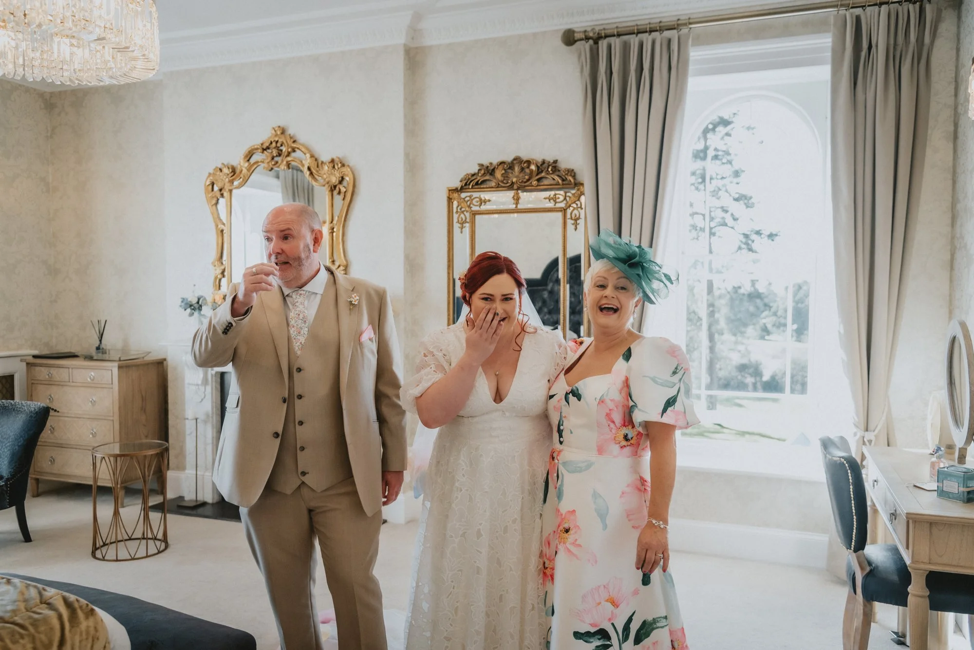 Three people in wedding attire standing in a beautifully decorated room with large window and elegant furniture, all smiling and laughing.