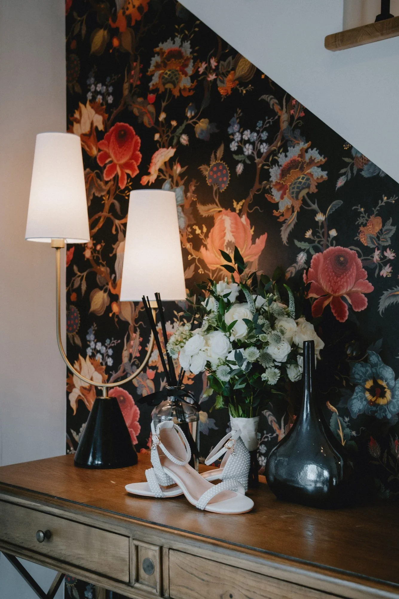 A wooden table with a pair of white, embellished high-heeled sandals, a black vase, a glass jar with black reed diffusers, and a bouquet of white flowers. Behind the table is a black wall with large pink, red, and purple floral wallpaper, and a white