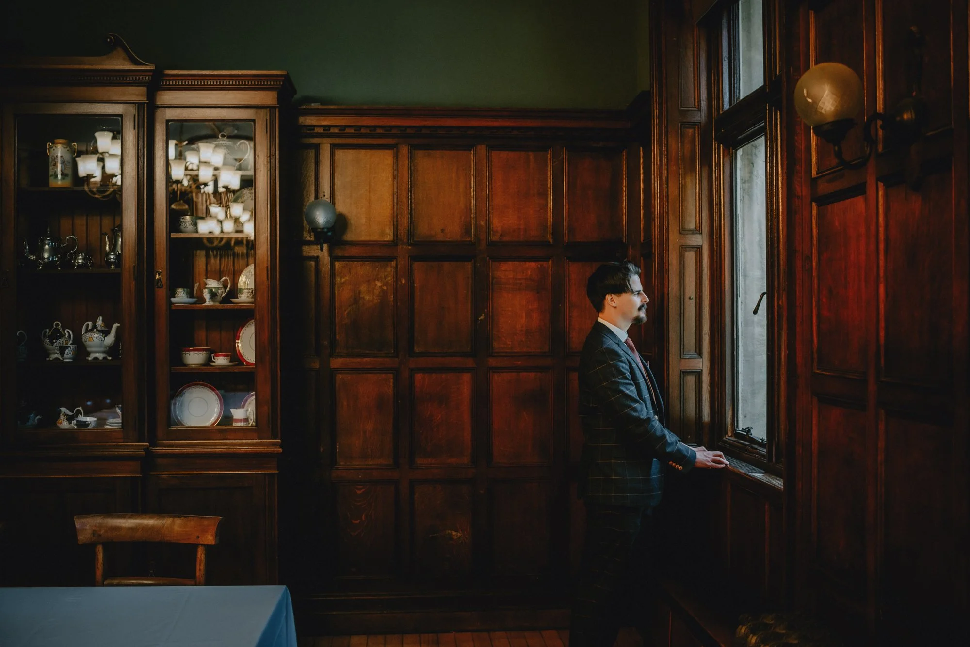 A man in a dark plaid suit stands at a window in a dimly lit room with dark wood paneling. Behind him is a glass cabinet filled with fine china and silverware, and a round wall sconce is mounted on the wall.