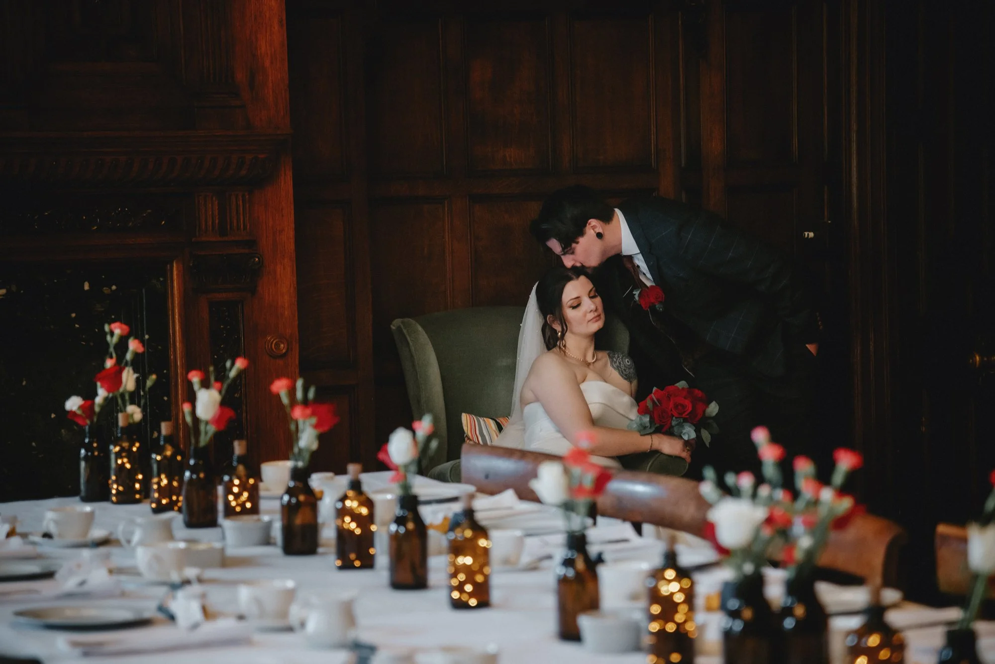 A bride in a white dress with jewelry, sitting on a chair holding a bouquet of red roses, with a groom leaning over her, in a wood-paneled room decorated for a wedding with a table set with flowers and candles.