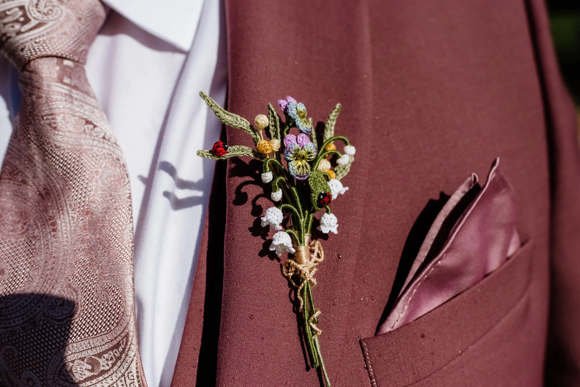 Close-up of a burgundy suit jacket with a embroidered flower boutonniere and a pink pocket square, worn with a white shirt and a patterned tie.