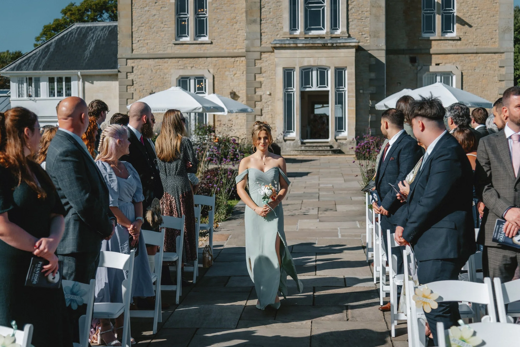 A wedding ceremony taking place outdoors in front of a historic building with a bride walking down the aisle, holding a bouquet, as guests standing on either side watch.