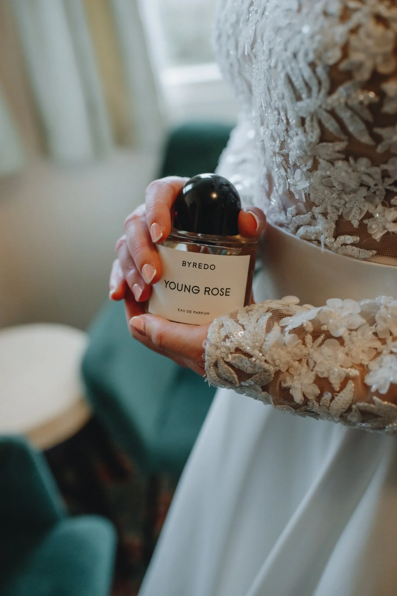 A bride in an embellished wedding dress holding a bottle of Byredo Young Rose perfume.