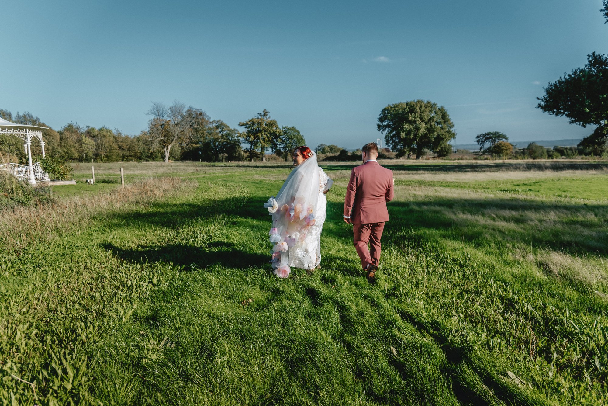 A bride and groom walking on a grassy field during their outdoor wedding, with trees and a blue sky in the background.