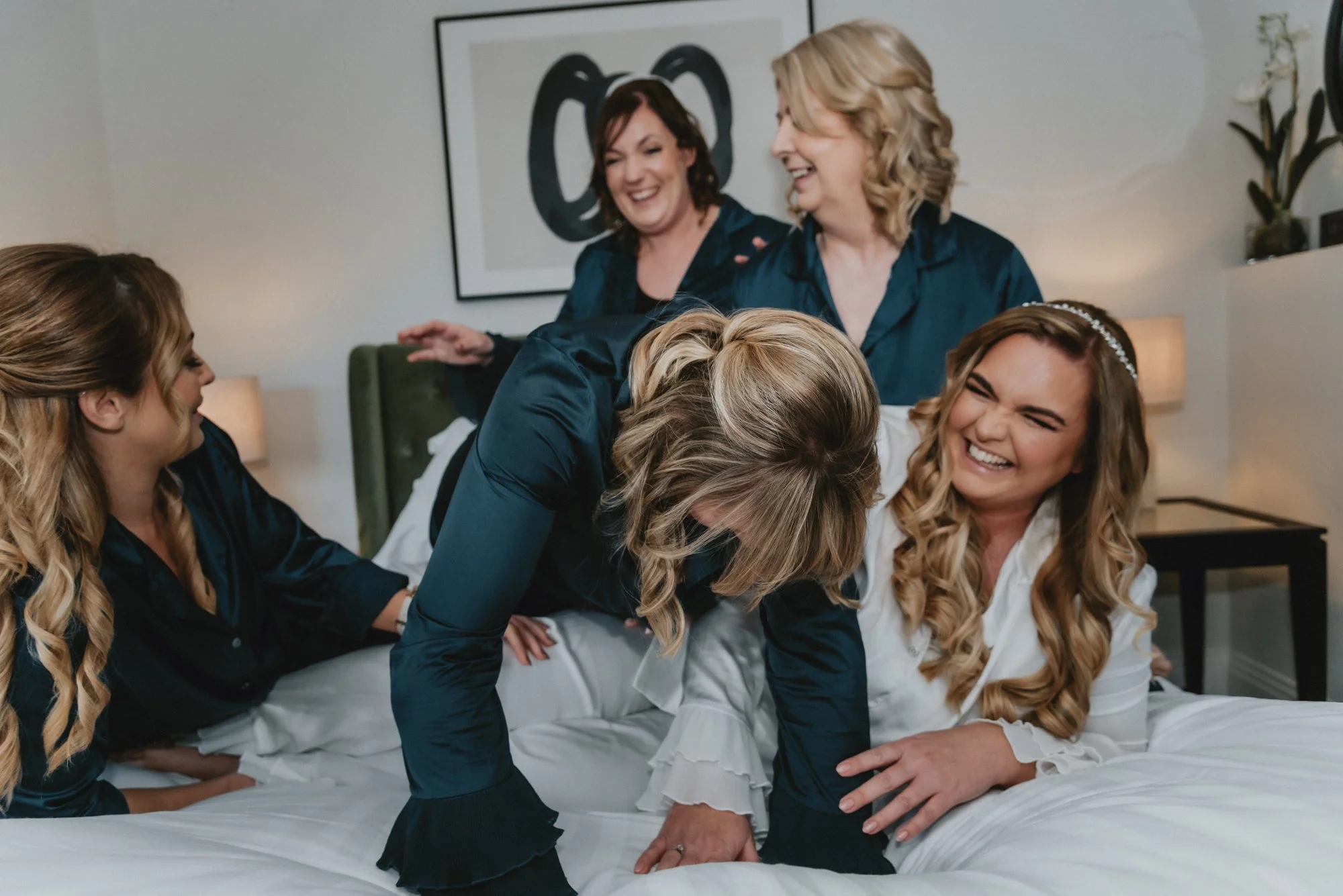 A group of women, likely a bride and her bridesmaids, joyfully laughing and playing on a bed in a bedroom with modern decor.