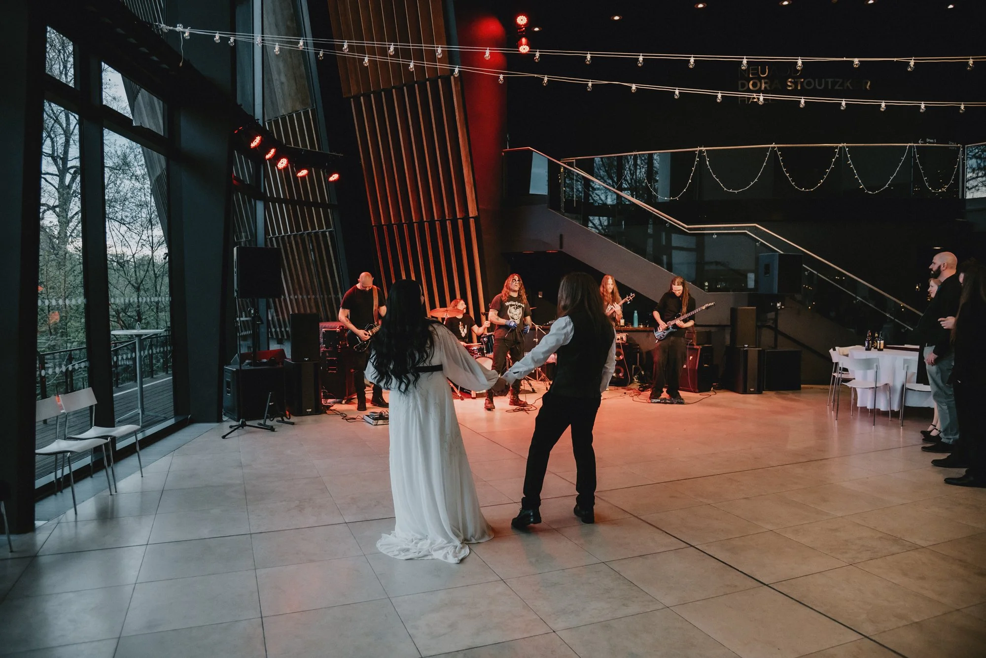 A bride and groom dance together in front of a live band playing on stage at a wedding reception in a modern venue with large windows and decorative string lights.