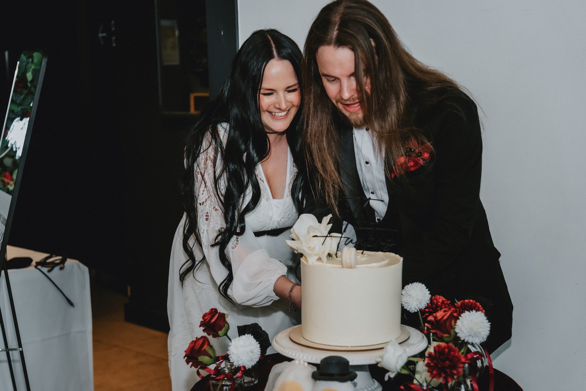 A woman and a man are cutting a wedding cake together, both smiling and looking down at the cake, surrounded by floral decorations.