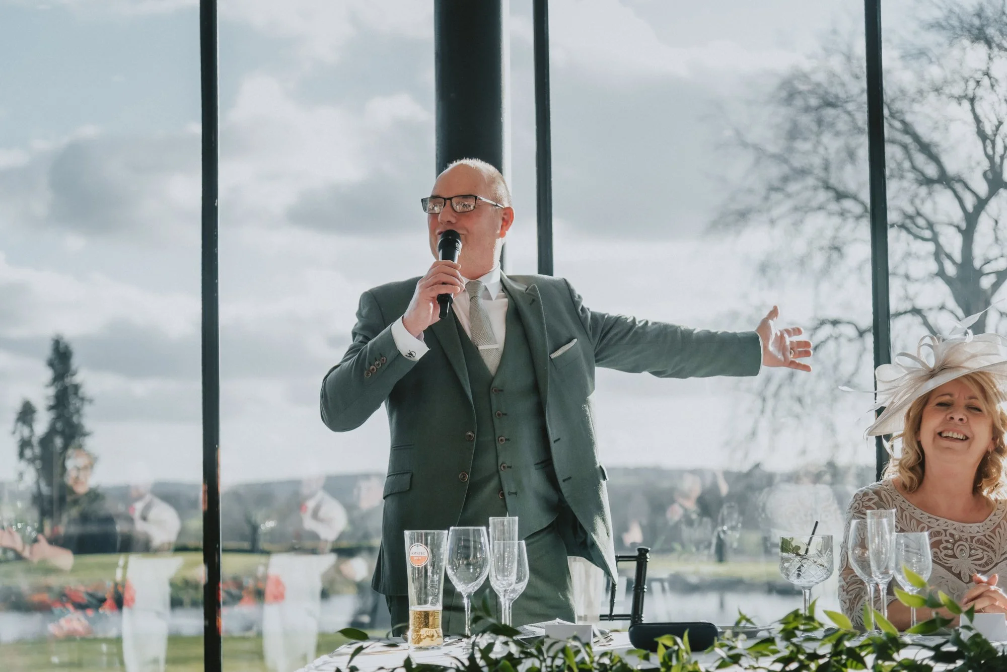 A man in a gray suit with glasses is speaking into a microphone at a wedding reception, with a woman in a white lace dress and wide hat smiling beside him. They are indoors near large windows showing an outdoor landscape with trees and a cloudy sky, 