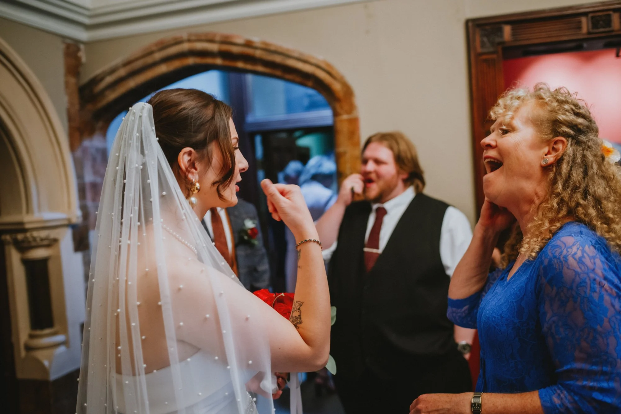 A bride with a veil and a necklace is smiling and holding a bouquet of red flowers, interacting with an older woman in a blue lace dress. Two men are in the background, one of whom is talking on the phone and wearing a vest and tie. The setting appea