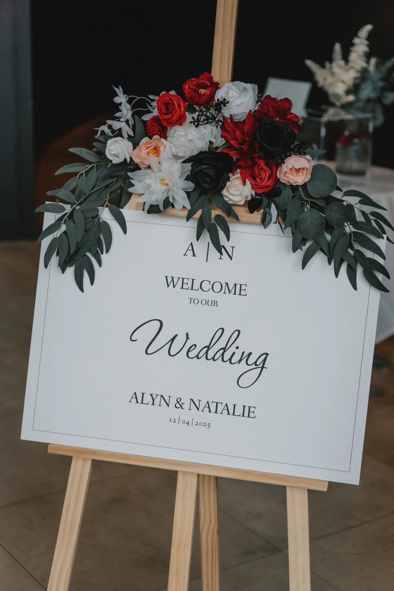 Wedding welcome sign with floral arrangement on wooden easel, featuring red, white, pink, black flowers, and greenery, for Alyn and Natalie on December 4, 2025.