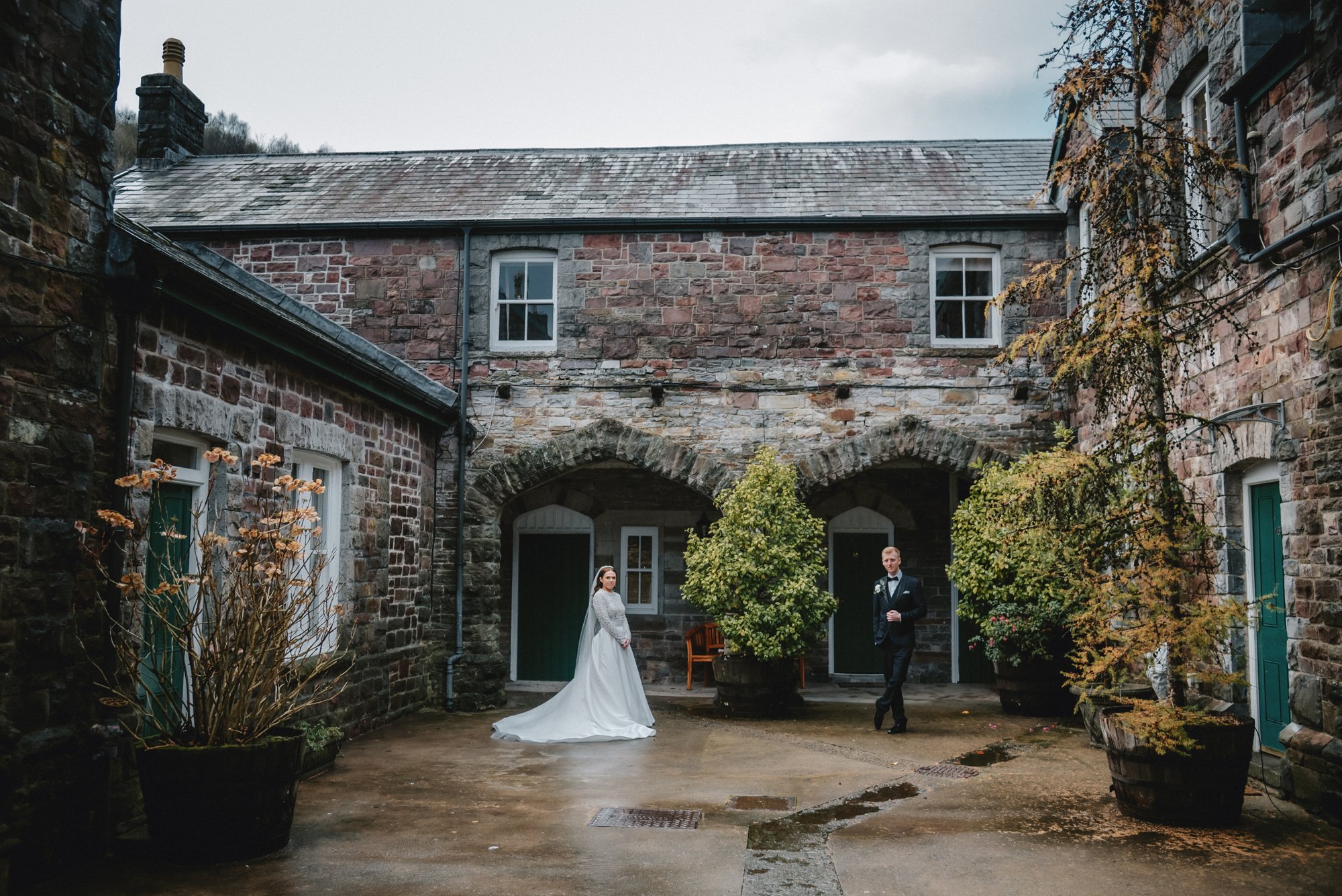 A bride in a white wedding gown and veil stands near two potted trees, while a groom in a tuxedo stands nearby in a courtyard with stone buildings, potted plants, and wet pavement.