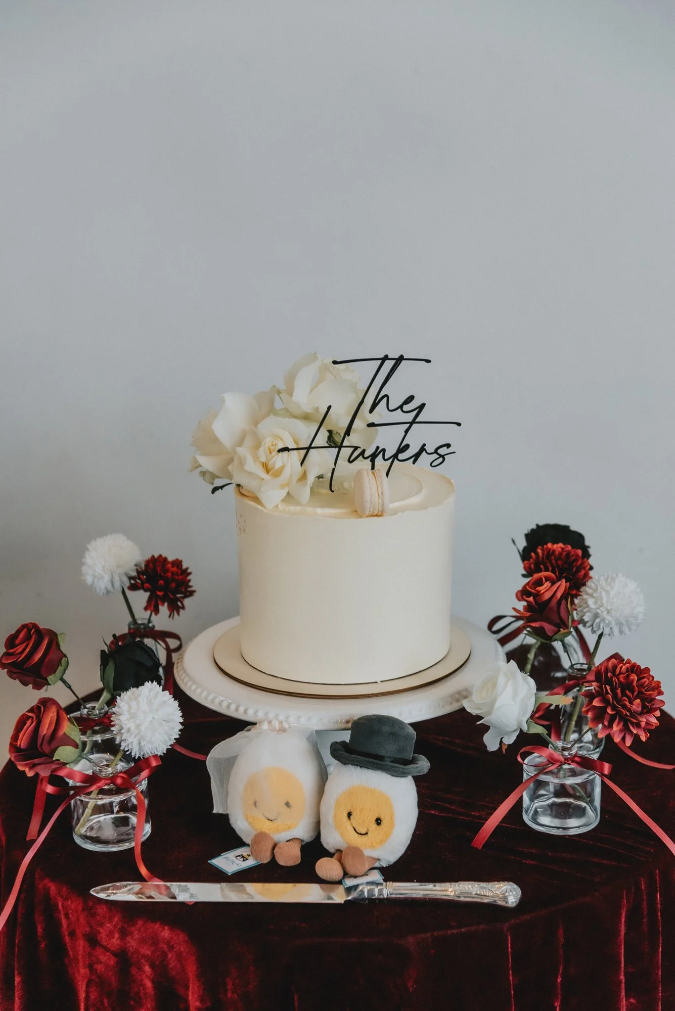 A wedding cake with white roses and a topper that reads 'They Talkers,' surrounded by red, white, and black flowers in glass vases on a velvet tablecloth, with plush doll toys and a cake knife in front.