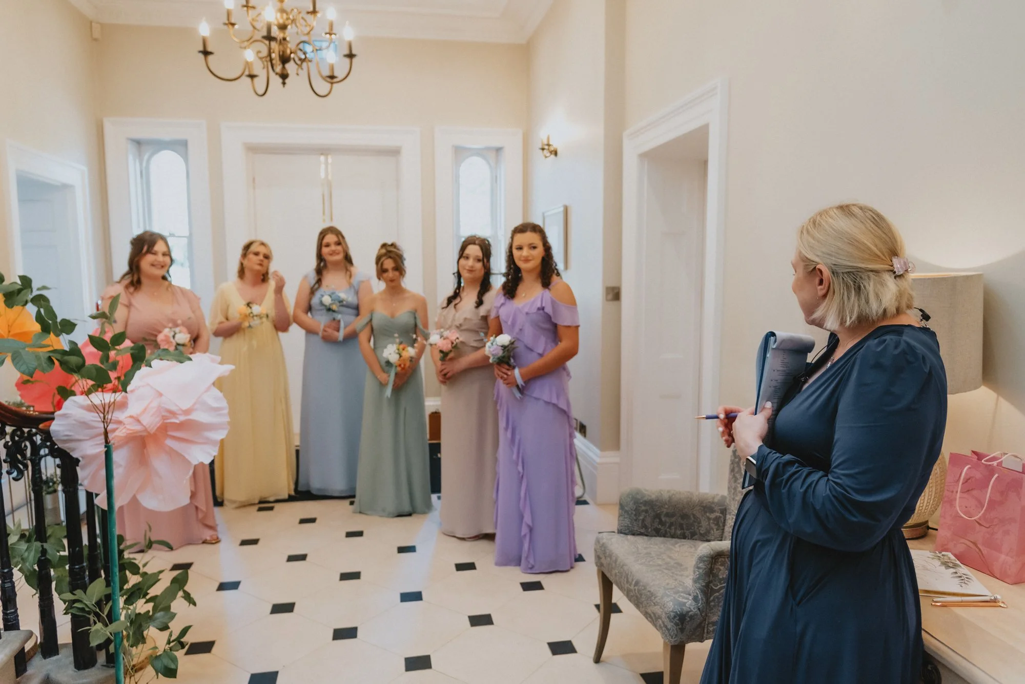 A woman giving a speech or speech notes to six bridesmaids standing in a line, holding bouquets, in a brightly lit, elegant room with chandeliers and white walls.