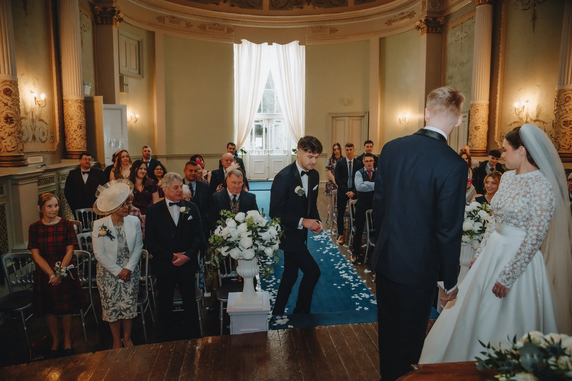 A wedding ceremony with a bride and groom standing at the altar, surrounded by guests inside an ornate, well-decorated hall.