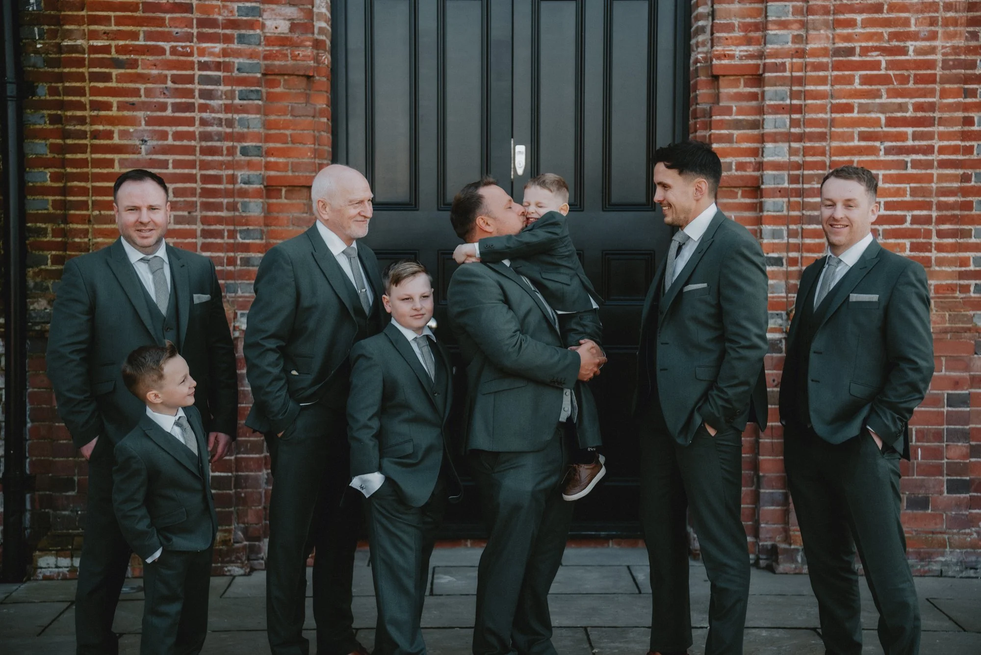 A group of eight men and boys in formal suits standing in front of a brick wall, with one man holding a young boy who is hugging him.